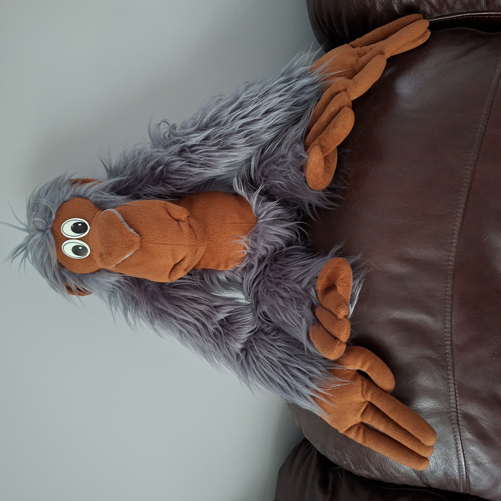 A plush toy orangutan with grey shaggy fur and orange hands and face sits on a brown leather sofa in front of a light grey wall. The orangutan is positioned centrally, its arms spread wide and resting on the back of the sofa. Its eyes are large and white with black pupils, and its nose is a prominent orange triangle. The sofa appears worn, with visible stitching and creases in the leather. The lighting is even, suggesting an indoor setting. There is no indication of the location being in Anapskaya, Russia or any specific city.