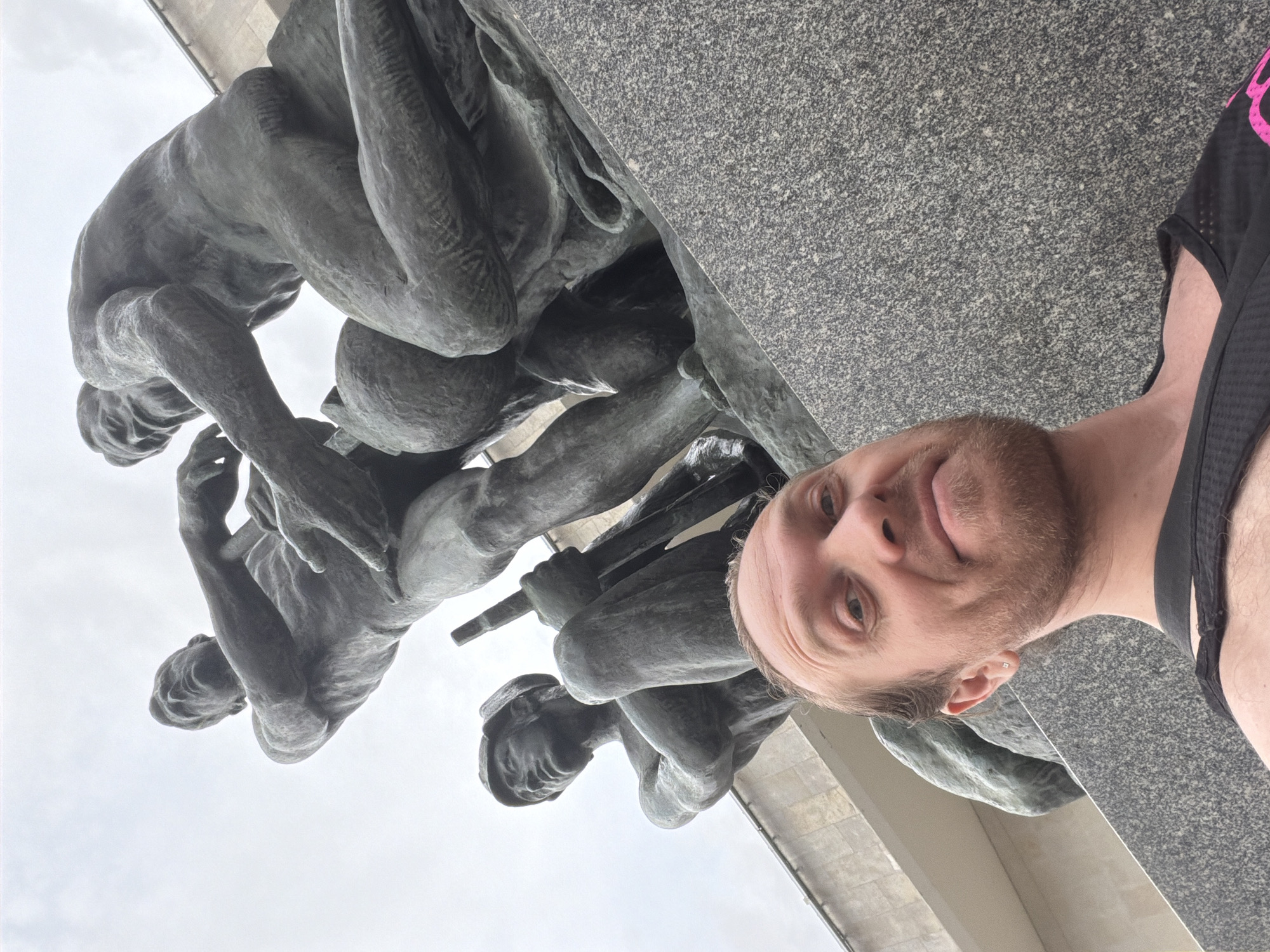 A low-angle, eye-level selfie captures a man with light brown hair and a beard, wearing a dark tank top, smiling faintly into the camera. He occupies the lower half of the frame, positioned in front of a monumental bronze sculptural group. The statues, rendered in a classical, heroic style, depict three muscular male figures on a textured grey granite base. To the left, a figure wearing a cap is seated, holding a long, slender tool (possibly a pickaxe or pole) resting on his shoulder, with his arms crossed on his chest. To the right, two figures stand or lean; one holds a large sphere or globe, while the other reaches towards it with an outstretched arm. The statues are set against an overcast sky and a light-colored building in the background. This monument is identified as part of the Monument to the Heroic Defenders of Leningrad at Victory Square (Ploshchad Pobedy) in Saint Petersburg, Russia.