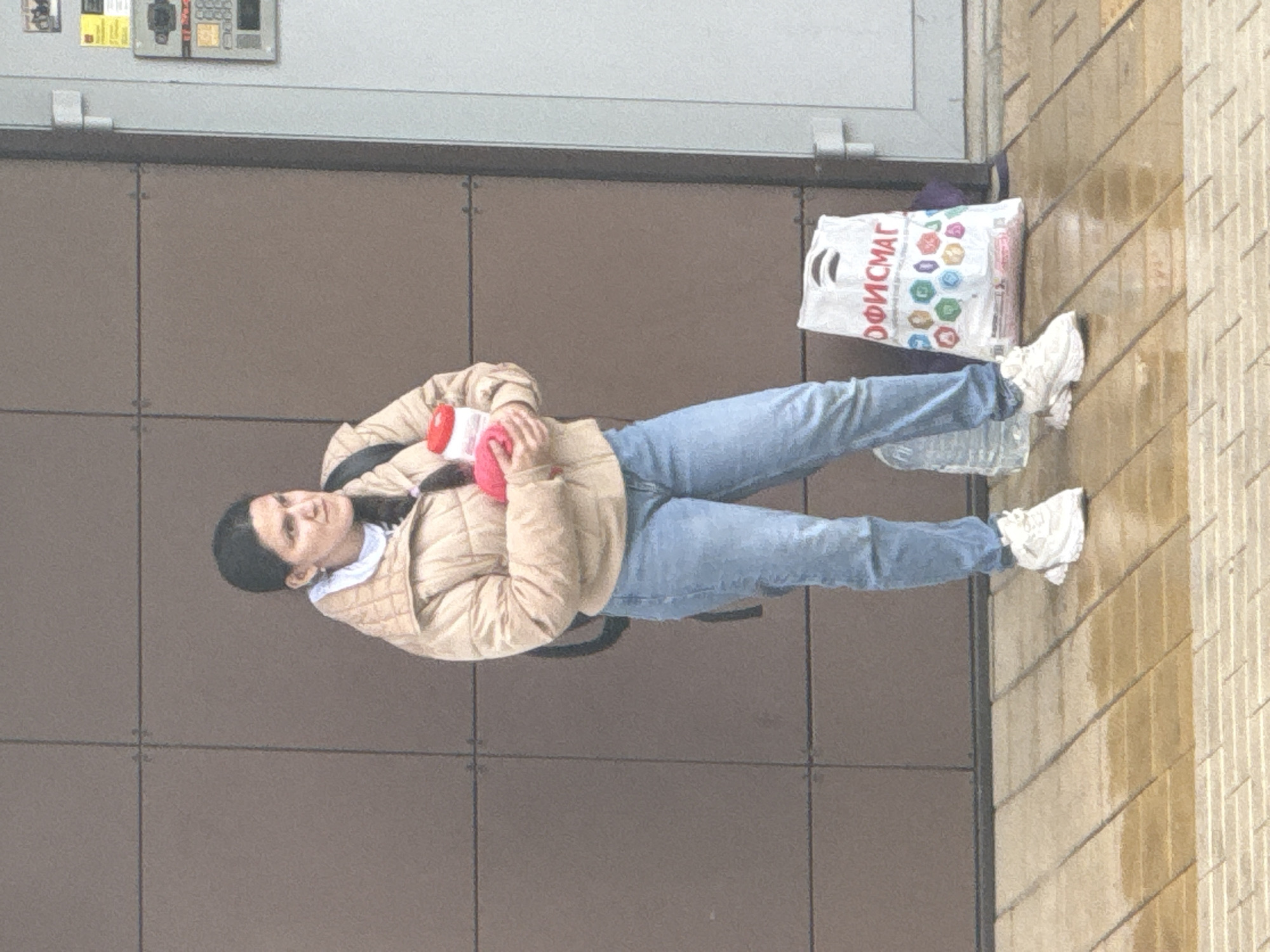 A woman stands on a sidewalk in what appears to be Rostov-on-Don, Russia, based on the Cyrillic text "ОФИСМАГ" on the shopping bag next to her. She is wearing a beige quilted jacket, light blue jeans, and white sneakers. She holds a red and white cylindrical object in her hands. A shopping bag and a plastic bottle are on the sidewalk beside her. She is standing next to a wall with a dark gray tiled surface, possibly a building facade, and a wet tile sidewalk.