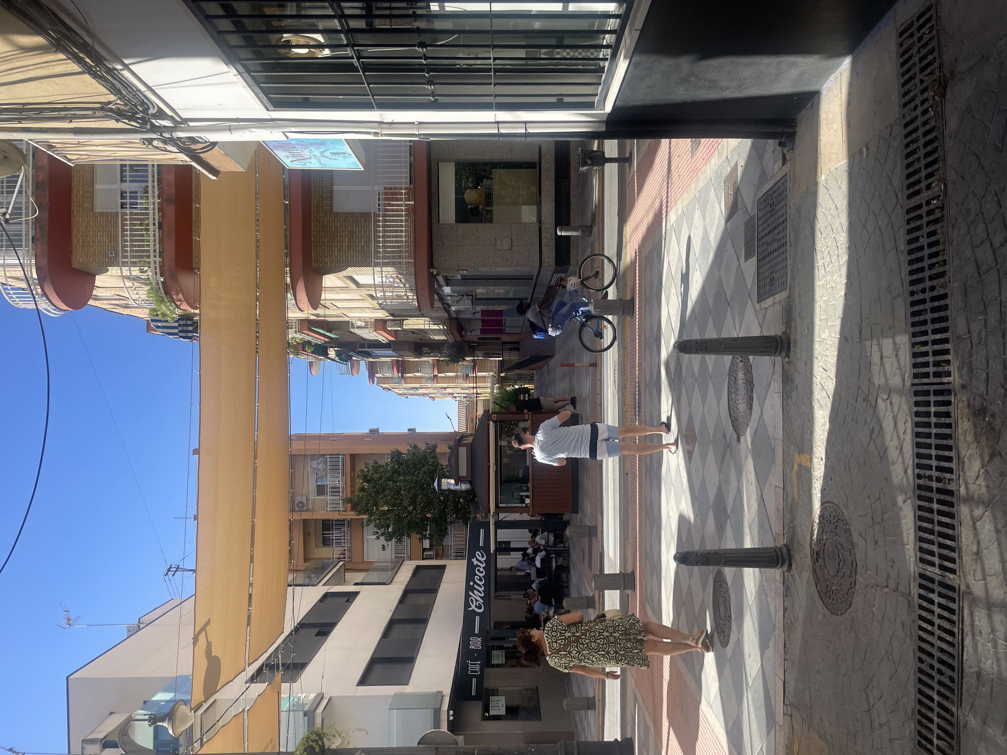 The image captures a sunlit street scene in Atarfe, Spain. A woman in a patterned dress walks away from the camera on a sidewalk paved with diamond-shaped tiles and darker gray stones. She carries a light-colored purse. Ahead of her, a man in a light blue t-shirt and striped shorts walks towards the background, also on the diamond-tiled sidewalk. A bicycle rider in a dark t-shirt and blue jeans is visible further down the street on the right, riding a light blue bicycle.
The street is lined with buildings on both sides. The building on the left features a cafe-bar with a prominent sign that reads "CAFÉ - BAR Chicote." Patrons are seated at tables outside the cafe. The buildings across the street have balconies with white railings and reddish-brown trim. A large, light-colored awning stretches across the street, casting shadows. The sky above is clear and blue.
The foreground of the image shows a section of the street with a drainage grate. In the middle ground, there are bollards along the edge of the sidewalk. The overall impression is a typical daytime scene in a Spanish town.