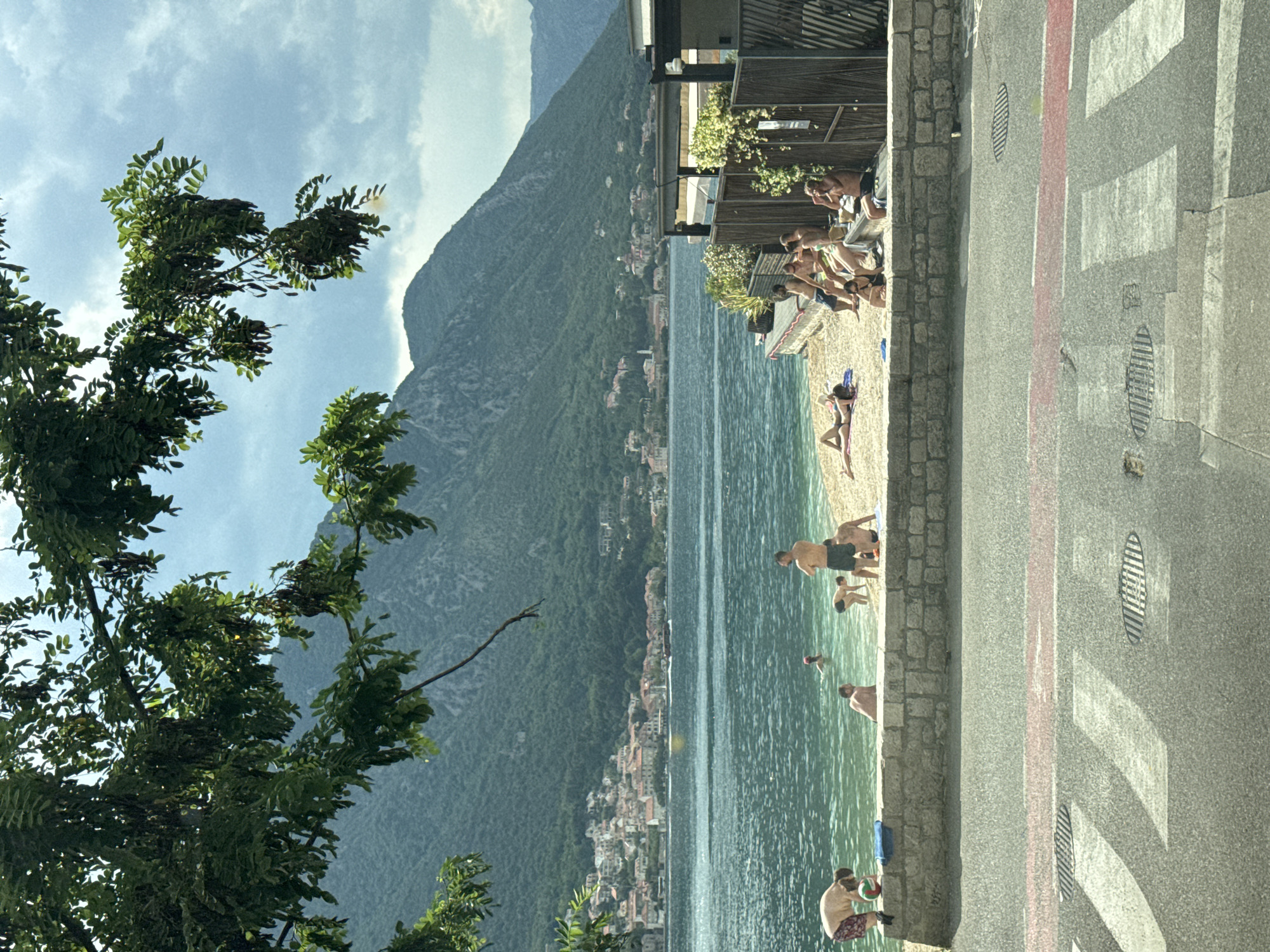 The scene is a beach area in Dobrota, Montenegro. A group of people are on the beach, some are swimming in the water, others are sunbathing or relaxing on the beach. A mountain looms in the background, and a tree is visible in the foreground. A road runs parallel to the beach, separated by a stone wall. There are crosswalk markings on the road. Some people are also gathered on an elevated patio area.