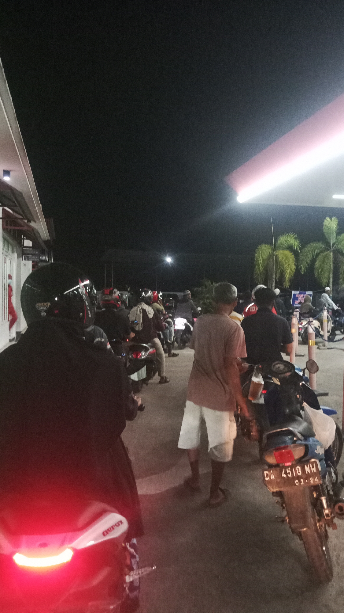 The image depicts an outdoor scene at night, likely at a gas station in Sungai Sipai, Indonesia. Several individuals are present, mostly on or around motorcycles. The foreground shows the rear of a motorcycle with its red taillight illuminated. To the right, a man in white shorts and a t-shirt stands near a motorcycle with a license plate reading "DA 4518 NW 03-24". In the background, more people and motorcycles are visible, suggesting a queue or gathering. Palm trees can be seen, and a brightly lit canopy with pink and white lighting overhead is present, characteristic of a gas station. The ground appears to be paved. The overall impression is a common nighttime scene of people refueling or waiting at a gas station.