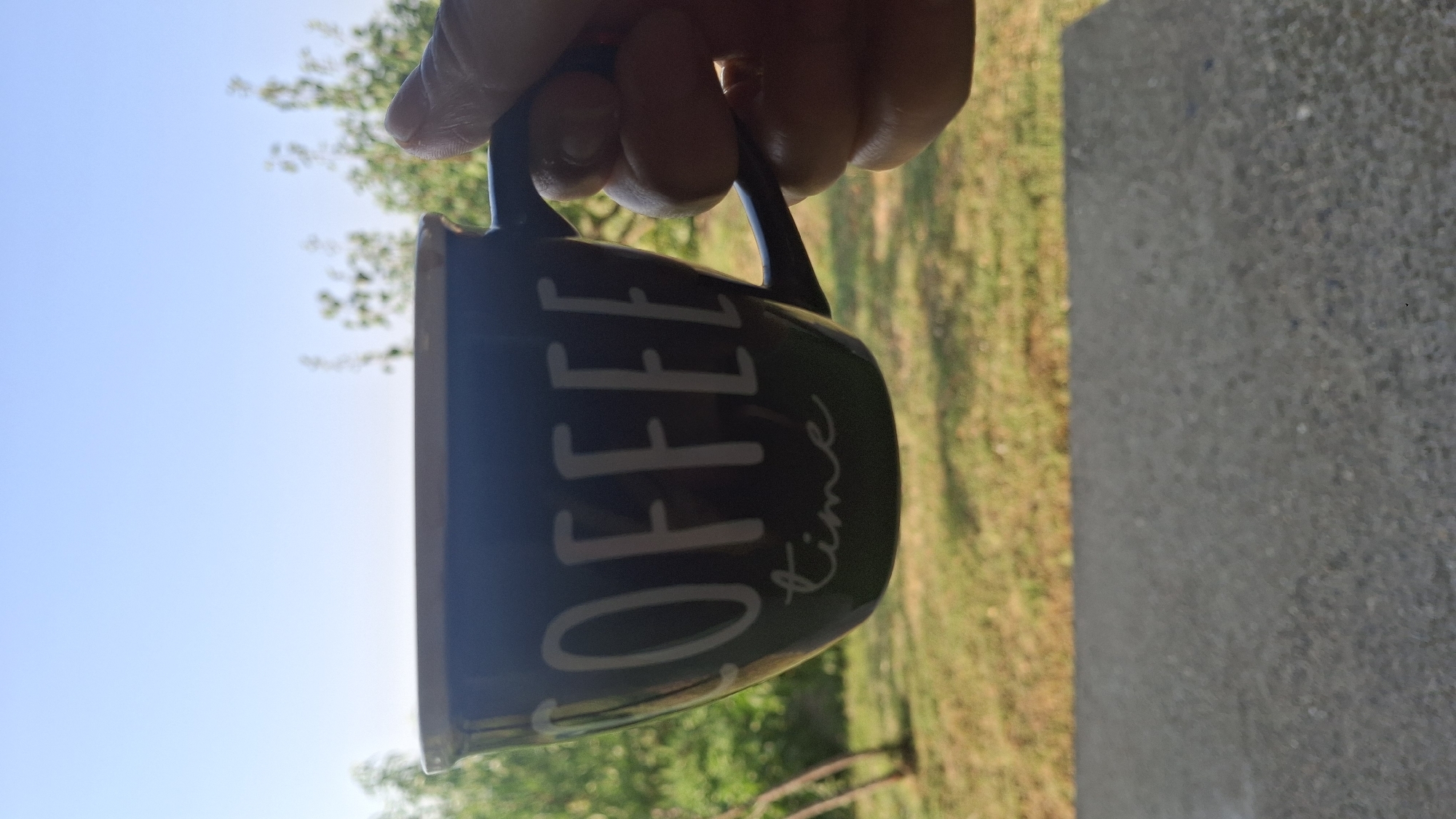 A person's hand is holding a dark blue mug with "COFFEE time" written in white script on it. The mug is positioned in the foreground. In the background, there is a blurred view of green foliage and a clear blue sky. The setting appears to be outdoors, possibly in a backyard or park. There is no specific information about the city of Butan, Bulgaria, present in the image.