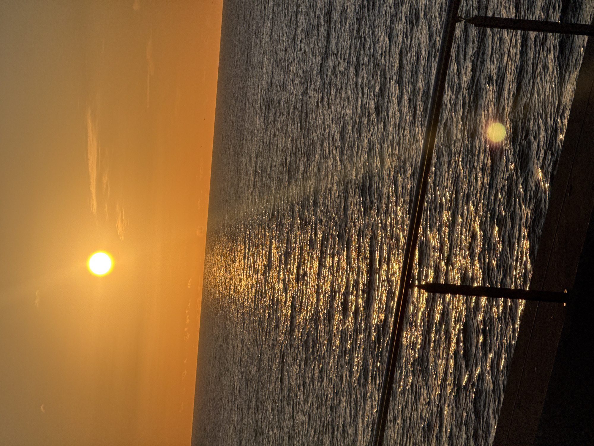 The image depicts a sunset over the sea, likely the Mediterranean, from a coastal location in Barcelona, Spain. The sun, a bright orb, is low on the horizon, casting a golden glow over the sky and reflecting on the rippling water surface. A dark structure, possibly part of a pier or boardwalk, is visible in the foreground, with vertical posts extending upwards. The sea's surface displays an interplay of dark and light, reflecting the setting sun.