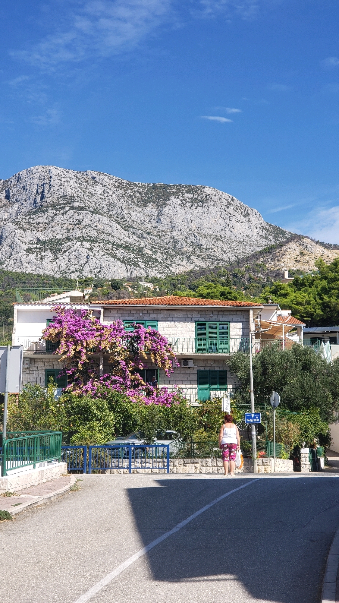 A woman stands on a street corner in Općina Tučepi, Croatia, facing away from the camera. She is wearing a white tank top and patterned capri pants. She is holding a white plastic bag in her right hand. In the background, a two-story building with white brick siding and green shutters is adorned with vibrant purple bougainvillea. A steep, rocky mountain looms behind the building, with green trees and some residential structures dotting its lower slopes. A street sign with the name "Podgradac" is visible on a pole near the woman. The sky is a clear, bright blue with a few wispy clouds. The street in the foreground is asphalt, with a white line marking a crosswalk. Shadows from unseen objects fall across the pavement.