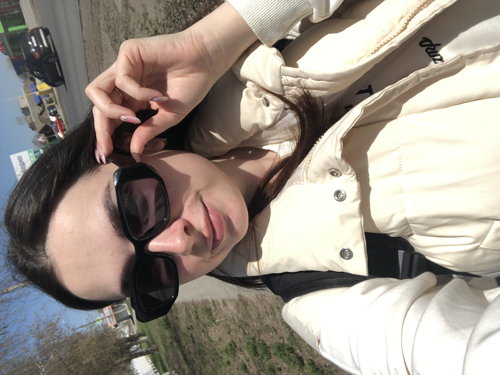 A woman is taking a selfie outdoors in what appears to be Chaikovsky, Russia. She is wearing a light-colored puffy jacket and black sunglasses, holding her hand near her face. In the background, there is a street with cars and buildings visible on a bright, sunny day. The ground is covered with grass and some bare trees.