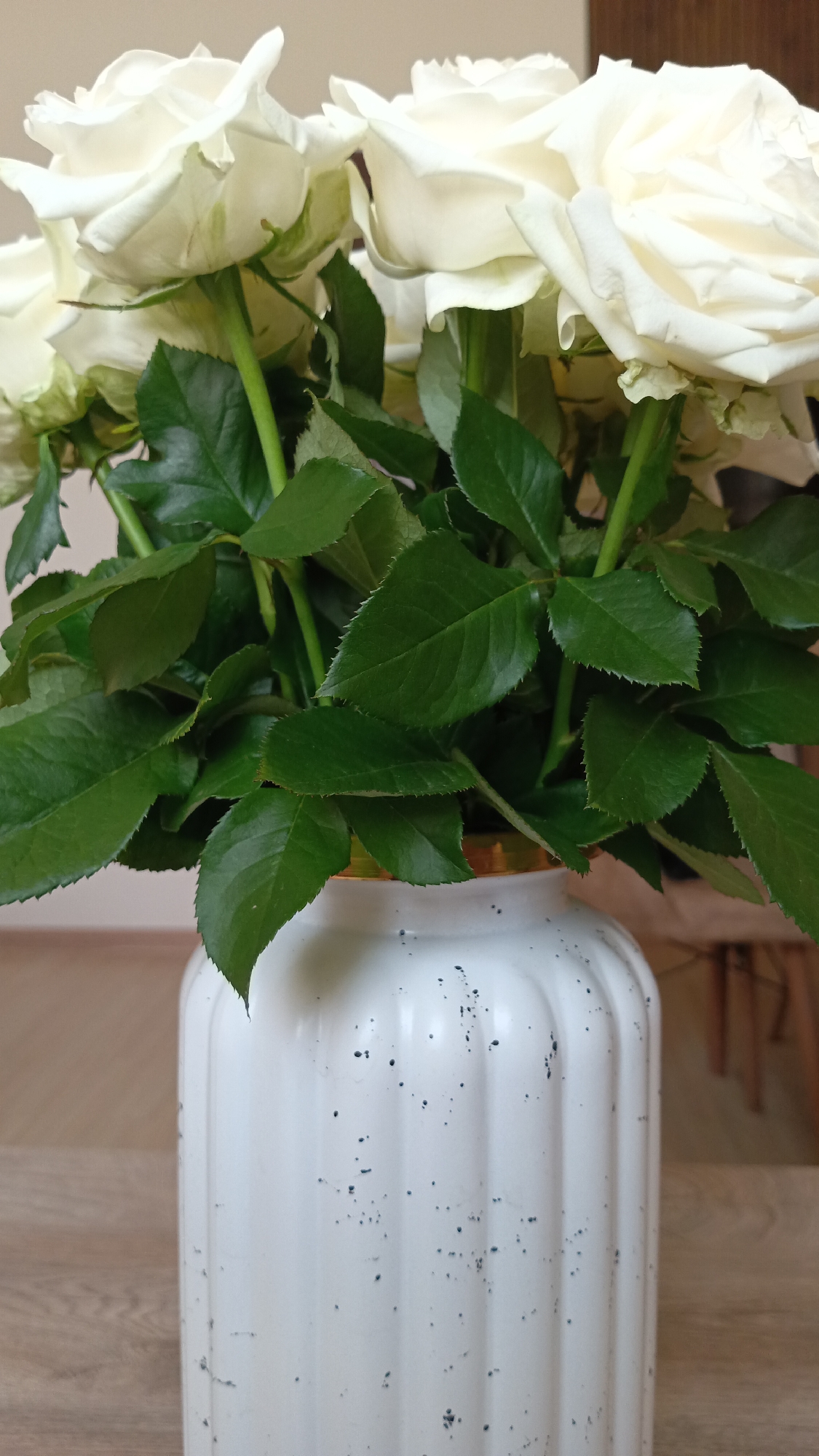 A close-up shot shows a bouquet of white roses in a fluted white ceramic vase with dark speckled accents. The vase has a gold rim. Lush green leaves surround the base of the roses. The background is out of focus, with a suggestion of a wooden table and furniture. The scene is indoors, with neutral-toned walls. The image does not provide any information about the city of Ashtarak, Armenia, or any people or actions.
