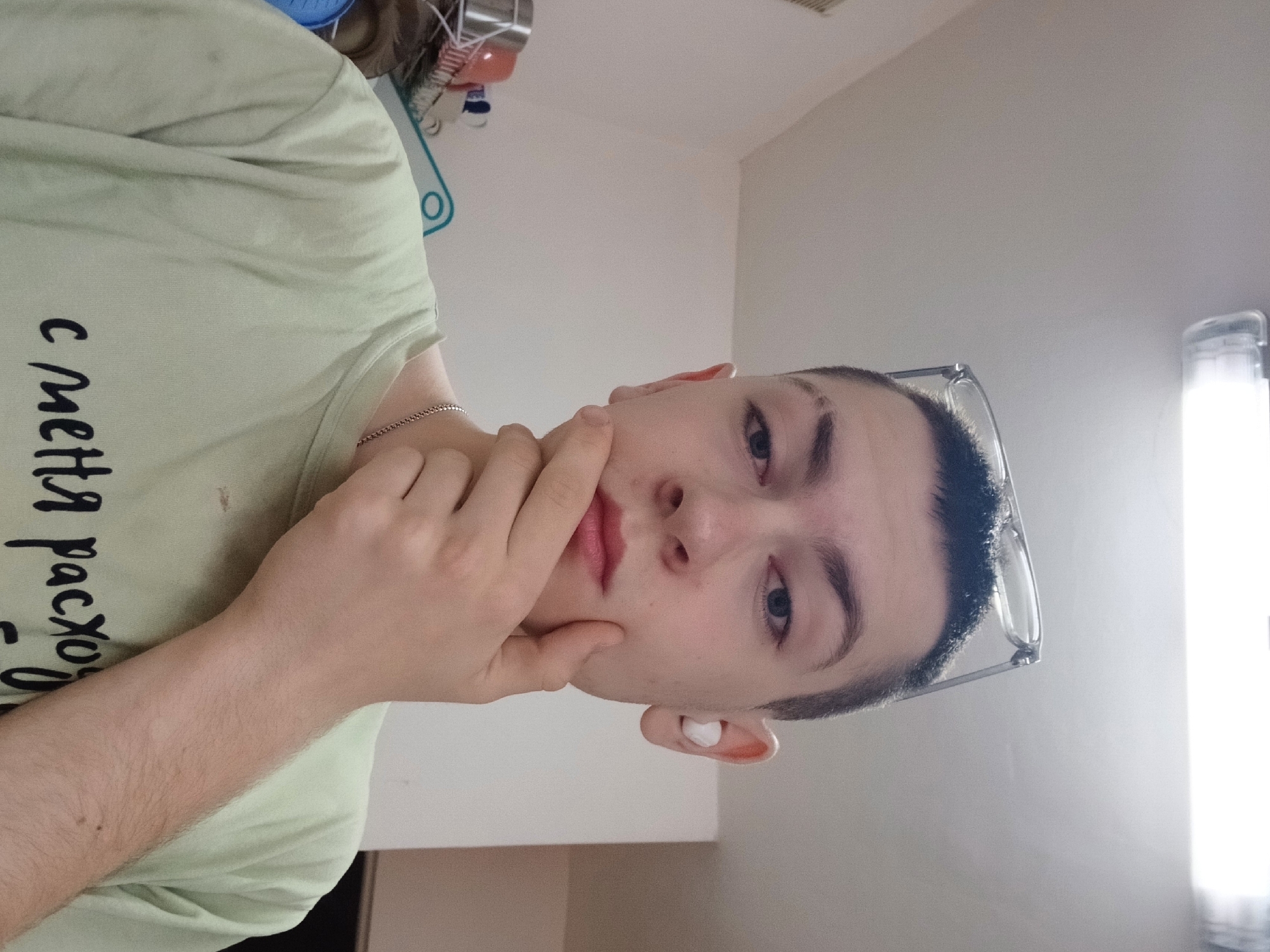 A young man is posing indoors. He is wearing a light green t-shirt with writing in Cyrillic. He is wearing glasses and a white earbud in his left ear. He is touching his chin with his hand. The background includes a wall and some household items. Based on the user's request, the location is Chaikovsky, Russia.