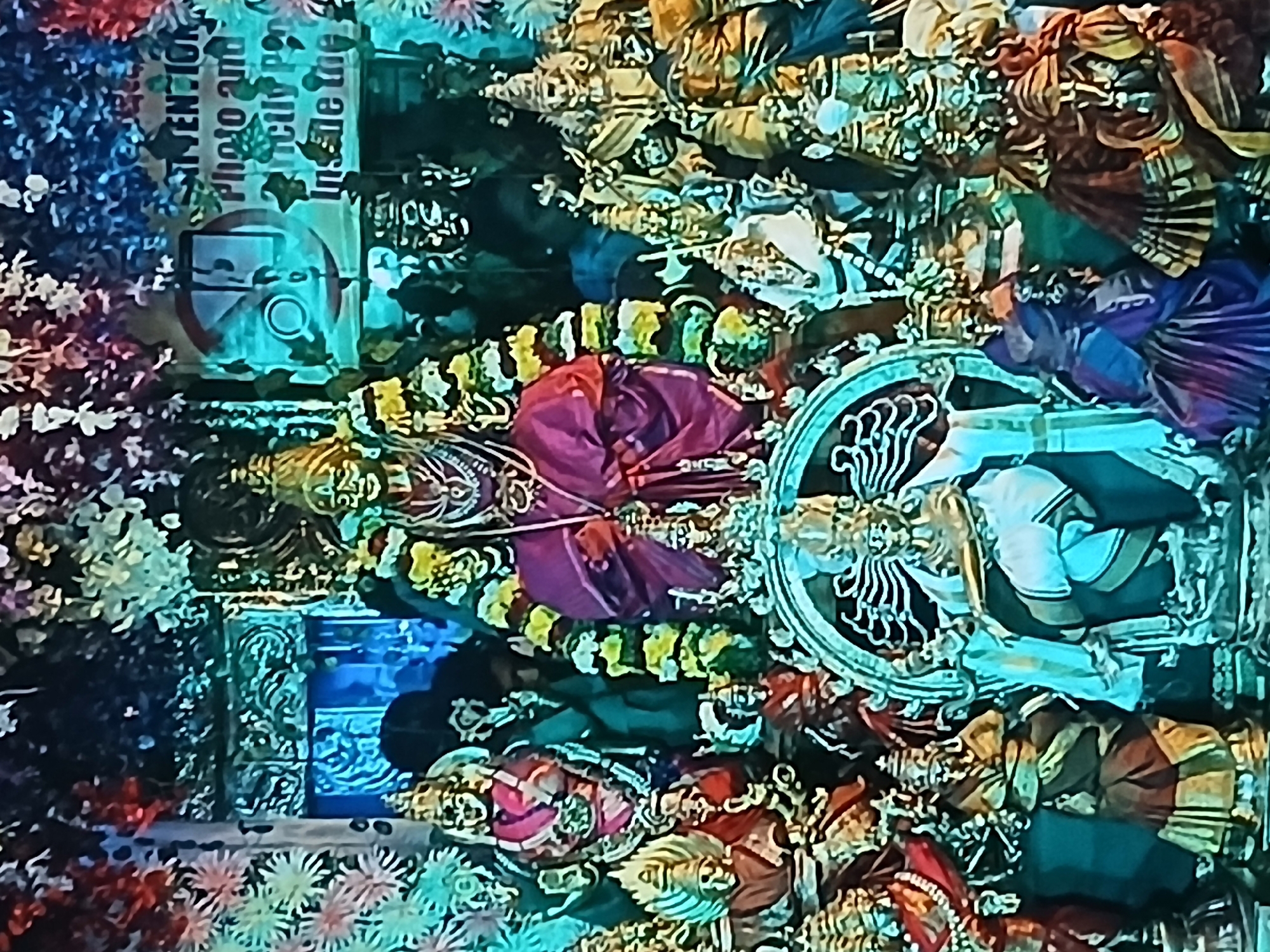 The image depicts a Hindu temple scene in Bengaluru, India. A sign indicates "Photo and Video Strictly Prohibited Inside the Temple", implying a restriction on photography. Several figures, adorned with garlands and traditional attire, are present. Two prominent figures are draped in a crimson robe and a white garment respectively, positioned in front of an ornate backdrop. The environment is decorated with an abundance of flowers, suggesting a festive or ceremonial occasion. The overall composition indicates a religious and cultural setting, possibly depicting deities or important religious figures.