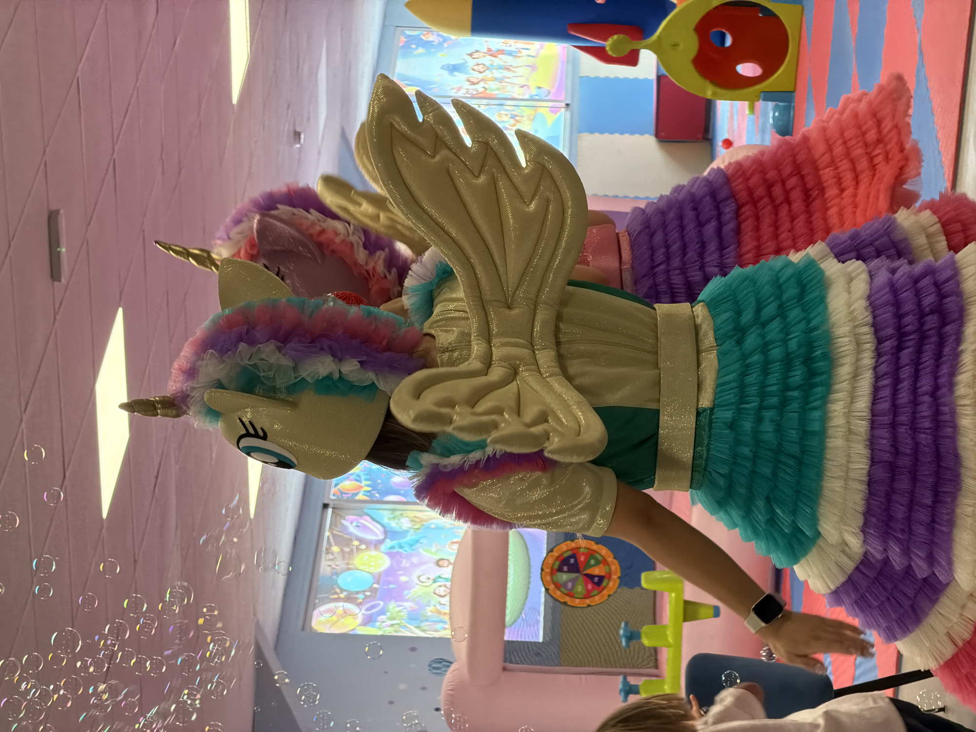 A person in a colorful unicorn costume with wings stands in what appears to be an indoor play area. The costume features a unicorn head with a golden horn and a rainbow-colored mane. The body of the costume is a shimmering gold with ruffled layers of fabric in teal, white, purple, and pink making up the skirt. Large, golden wings extend from the back of the costume. The setting includes padded, multi-colored floor mats and some play structures, including a yellow and red toy. In the background, there are colorful murals or posters on the walls, one depicting cartoon characters and another with what looks like underwater imagery. Bubbles are floating in the air in the foreground, suggesting an activity like bubble blowing is occurring. The lighting appears to be artificial, consistent with an indoor environment. The overall atmosphere seems festive and playful. The image is oriented vertically and taken from a slightly low angle.