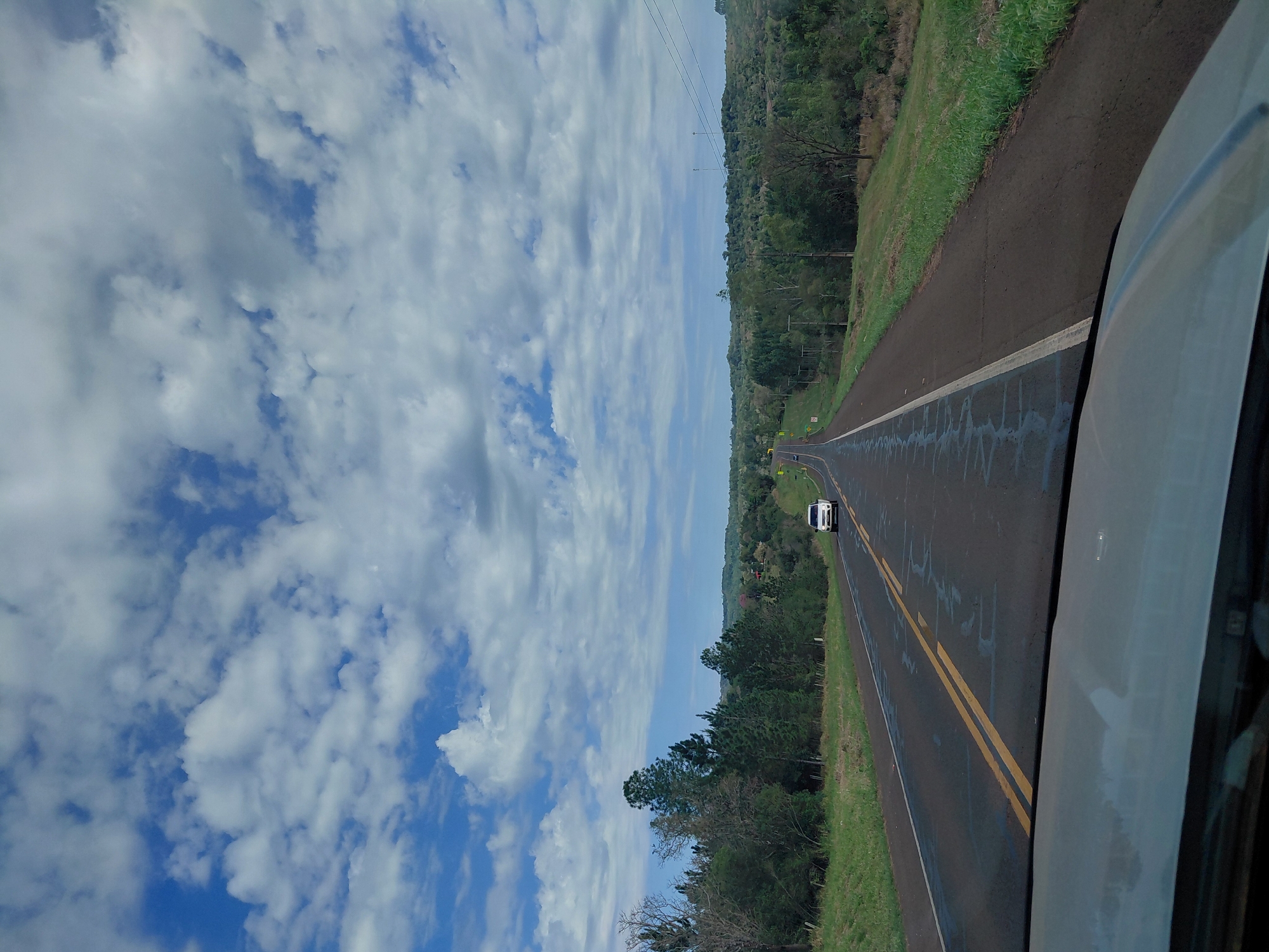 A white SUV is driving on a paved road that stretches into the distance, flanked by lush green vegetation and trees. The sky above is a vibrant blue with scattered white clouds. The road has double yellow lines in the center and a white line on the right side. The pavement appears wet in places, suggesting recent rain. The surrounding landscape is hilly and densely wooded. The image appears to be taken from the perspective of another vehicle, looking forward. The scene depicts a rural highway in Picada Boca, Paraguay.