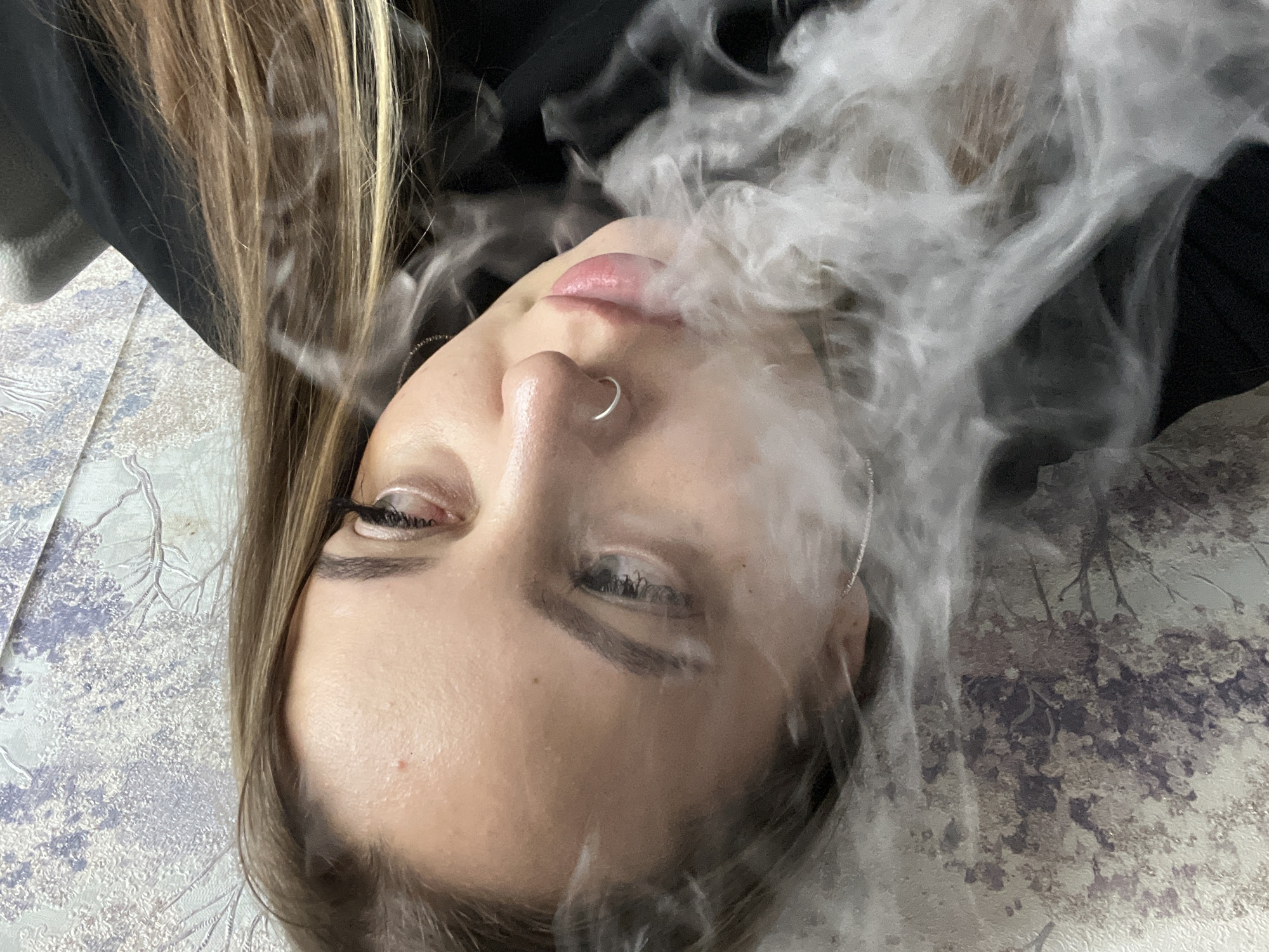 A woman with fair skin, light brown and blonde hair, and a silver nose ring is lying on a patterned surface. She appears to be exhaling smoke or vapor. The patterned surface appears to be a blanket or cloth. The location, Novopokrovskaya, Russia, is not discernable from the image.