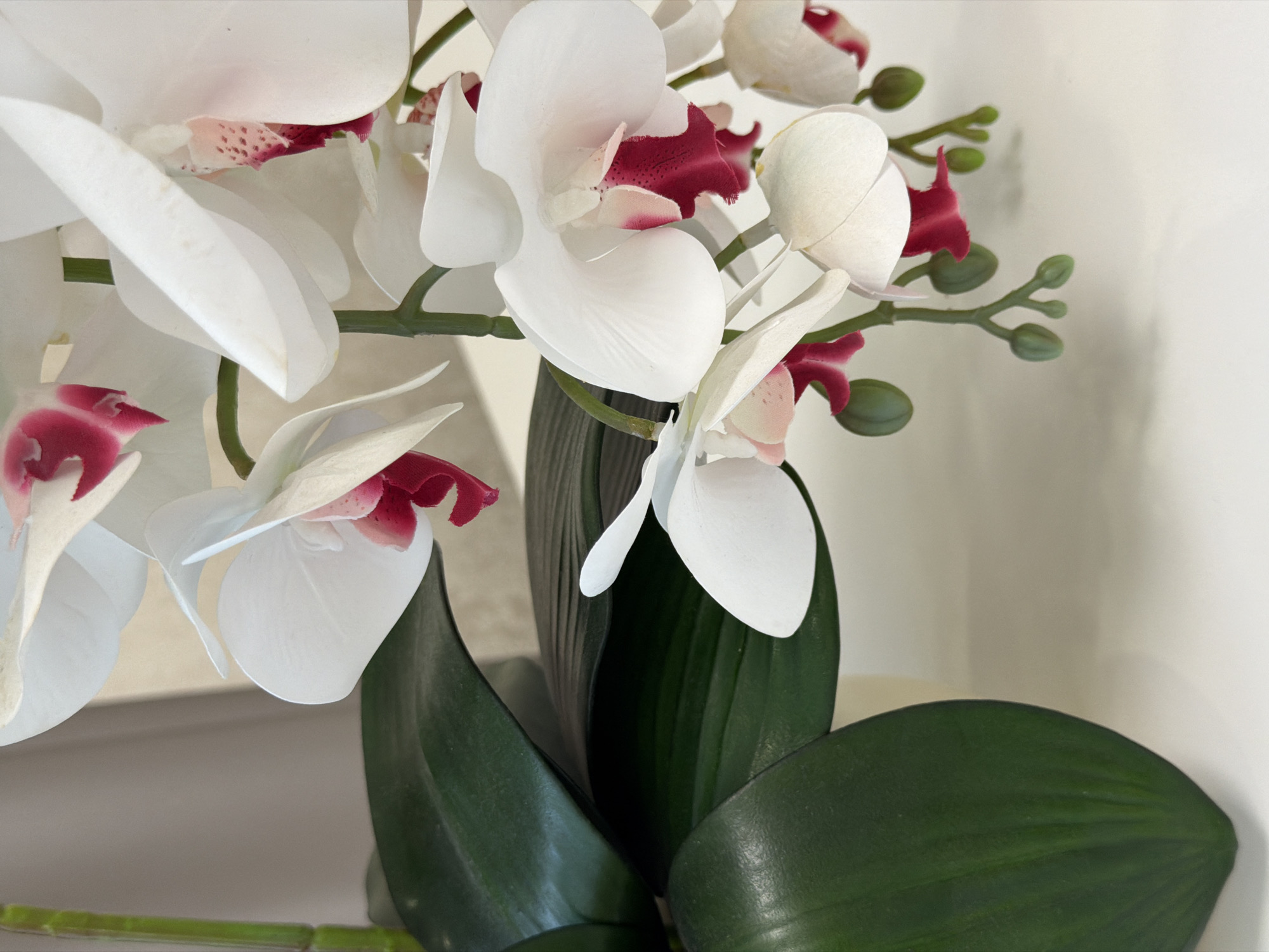 A close-up, eye-level shot features a cluster of white orchids with fuchsia centers, interspersed with deep green orchid leaves and thin green stems. The flowers are artificial, as indicated by their uniform texture and lack of natural imperfections. The orchids are arranged in a pot, partially visible at the bottom left, though the pot itself is not the focus. The background consists of a textured beige curtain and a white surface, suggesting an indoor setting, possibly a home or office. The lighting is soft and diffused, creating gentle shadows. There are no people or animals present. The scene is static and still.