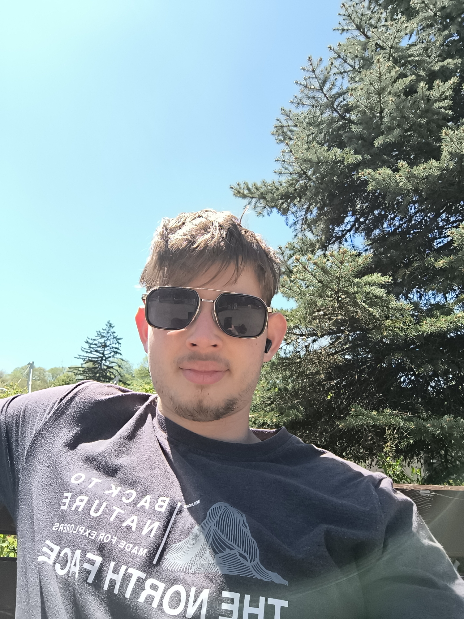 A selfie depicts a young man wearing sunglasses and a dark t-shirt with white lettering, sitting outdoors under a clear blue sky. A large pine tree is behind him to the right. The location is unspecified, and there is no indication that the location is Sofia, Bulgaria.