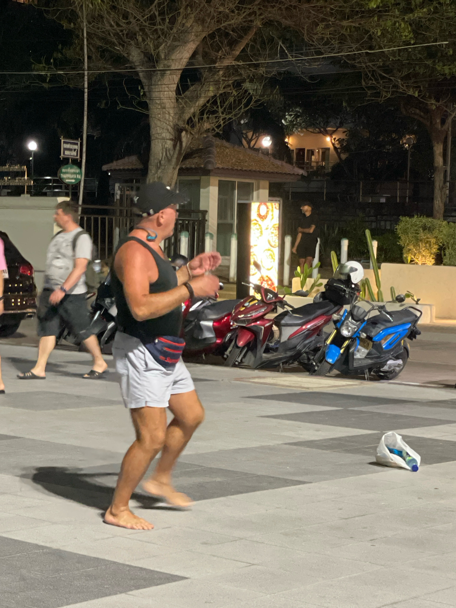 This image depicts a nighttime scene on a street in Pattaya City, Thailand, with a focus on a barefoot man in the foreground. The man, wearing a black tank top, grey shorts, a black baseball cap, and a red fanny pack, appears to be mid-stride or dancing. He has blue earbuds in his ears. To his left, another man in a grey t-shirt and black shorts walks away from the camera. In the background, several motorbikes are parked. Behind the motorbikes, a building with a thatched roof and a glass-fronted kiosk are visible, illuminated by interior lighting. Further back, trees with bare branches are prominent, and streetlights are casting a glow. A white plastic bag containing a blue bottle and other items lies on the ground in the lower right portion of the frame. The ground is paved with square tiles.