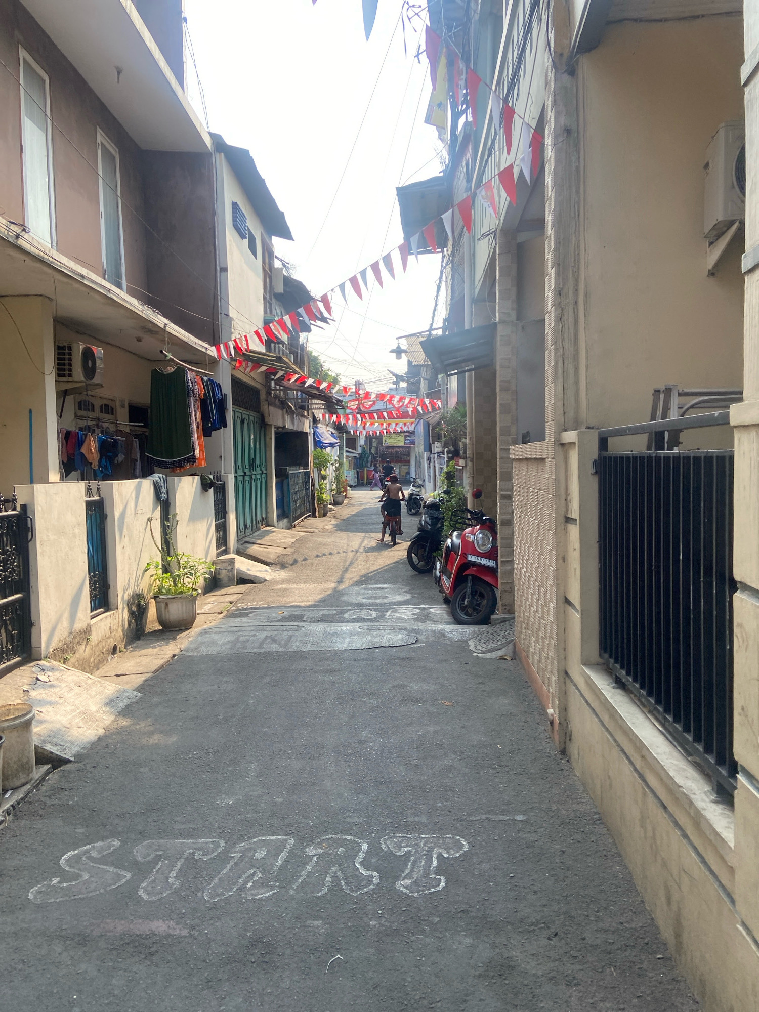 The image shows a narrow street in North Jakarta, Indonesia, lined with buildings and decorated with red and white triangular flags, suggesting a festive occasion. The street is paved and has "START" written in white chalk on the asphalt in the foreground. Several motorcycles are parked along the sides of the street, and a few people are visible in the distance, walking or riding motorcycles. Laundry is hung out to dry on a clothesline on the left side of the street. The overall impression is of a lively urban neighborhood.