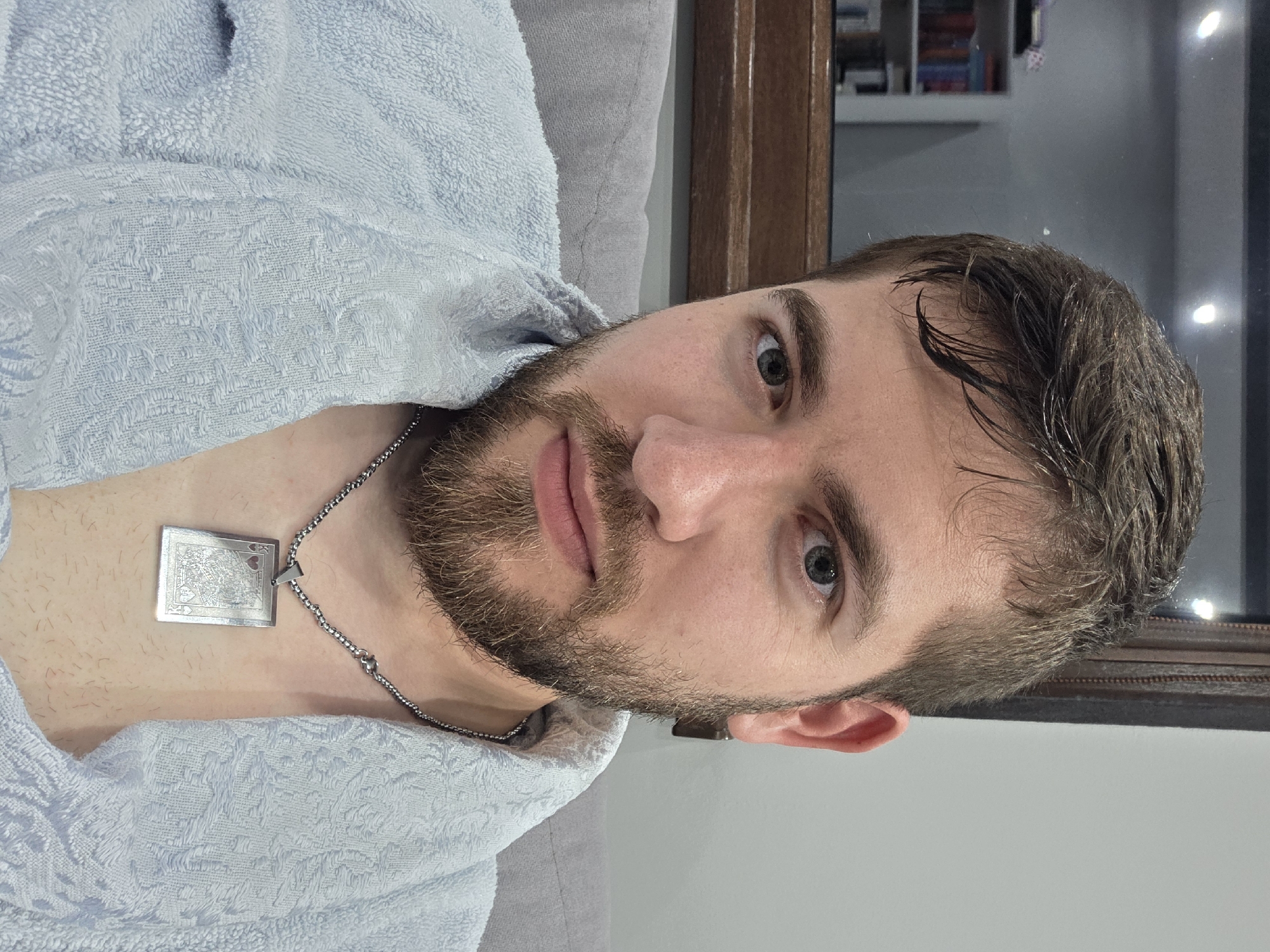 A man is positioned indoors, likely in a bathroom, wearing a blue bathrobe and a silver necklace with a card pendant. He has short brown hair, a beard, and fair skin. The background includes a gray surface, possibly a couch, and a wooden-framed mirror reflecting bookshelves and other indistinct items. The image orientation is sideways. There is no information indicating a location in Yagodovo, Bulgaria.