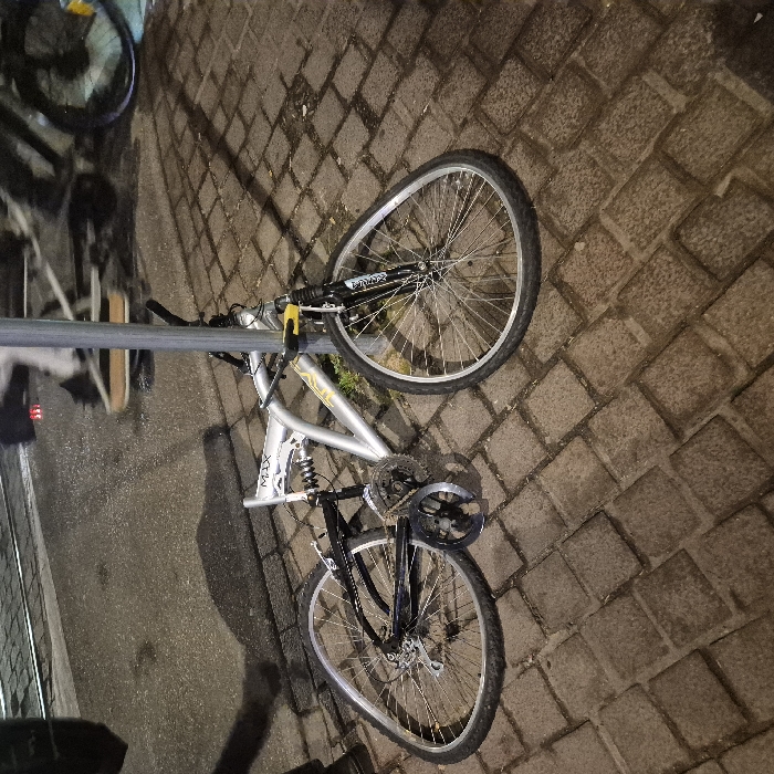 A silver and black mountain bike lies on its side on a brick pathway, seemingly secured to a gray metal pole with a yellow lock. The bike has front and rear suspension. Its front tire is fully inflated while the back tire appears deflated. Part of a second bicycle is visible in the upper left corner. The lighting suggests it's nighttime.