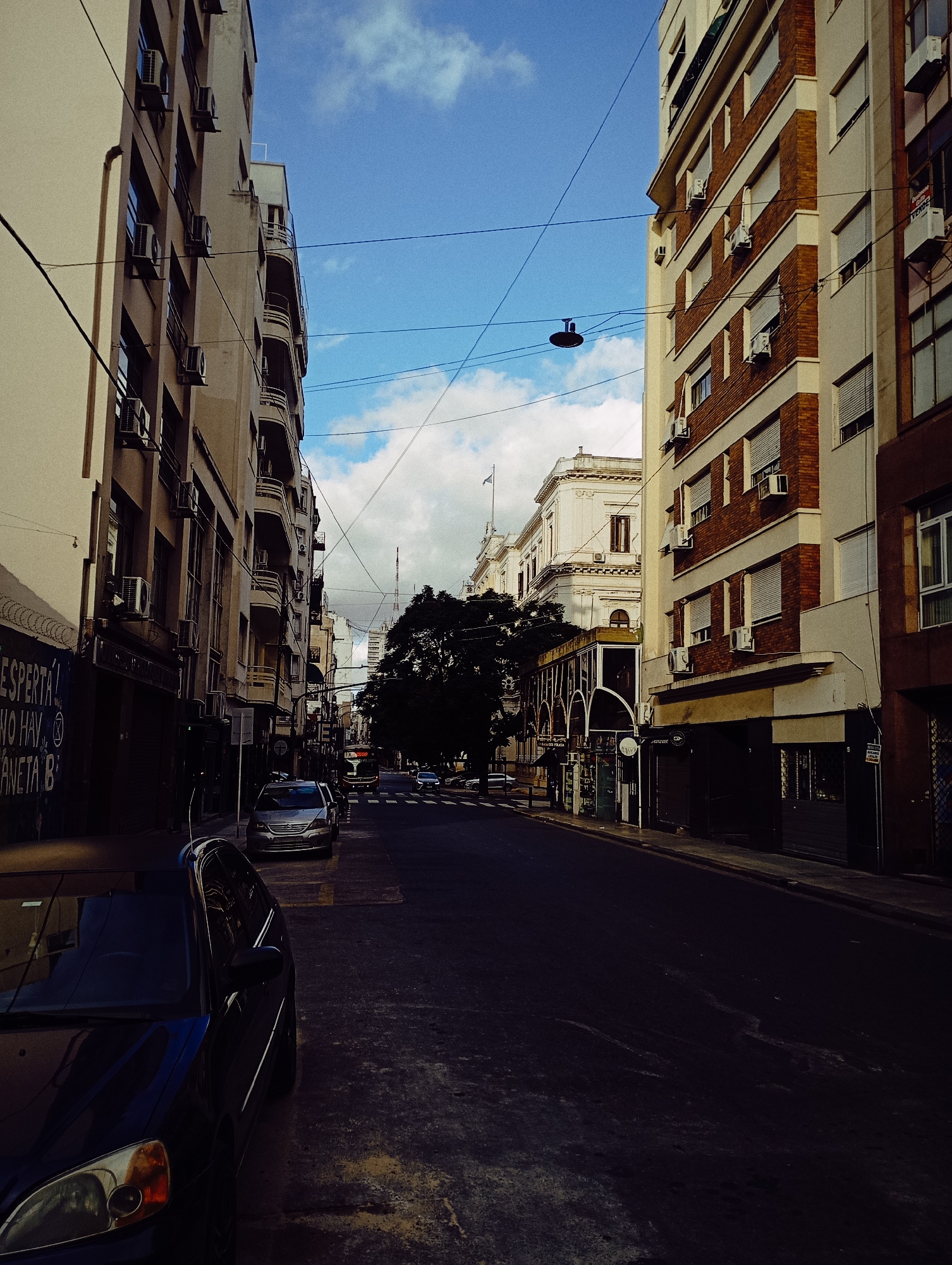 The image shows a street scene in Buenos Aires, Argentina. Buildings line both sides of the street, with a view of a government building visible at the end. A black car is parked on the left side of the street, and other cars are parked further down the street. Overhead wires cross the street. The street is paved and appears to be wet. The sky is blue with clouds. The area appears to be a commercial area, possibly downtown.