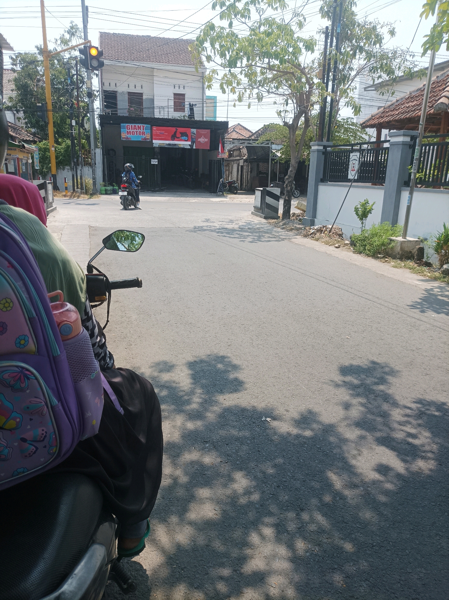 The image depicts a street scene in Sumuragung, Indonesia, on a sunny day. In the foreground, a person is seated on a motorcycle, with a purple backpack with butterfly and flower patterns visible. The motorcycle's handlebar and side mirror are also in view. The road is paved with asphalt and has tree shadows cast upon it. In the middle ground, a person on a motorcycle is stopped at a traffic light, which is displaying a yellow signal. The traffic light is mounted on a yellow pole. Adjacent to the traffic light is a building with a dark facade, featuring banners advertising "GIANT MOTOR" and a scooter. Further down the street, more buildings and a fence with a crest are visible. The environment appears to be a typical street in a populated area, with power lines overhead.