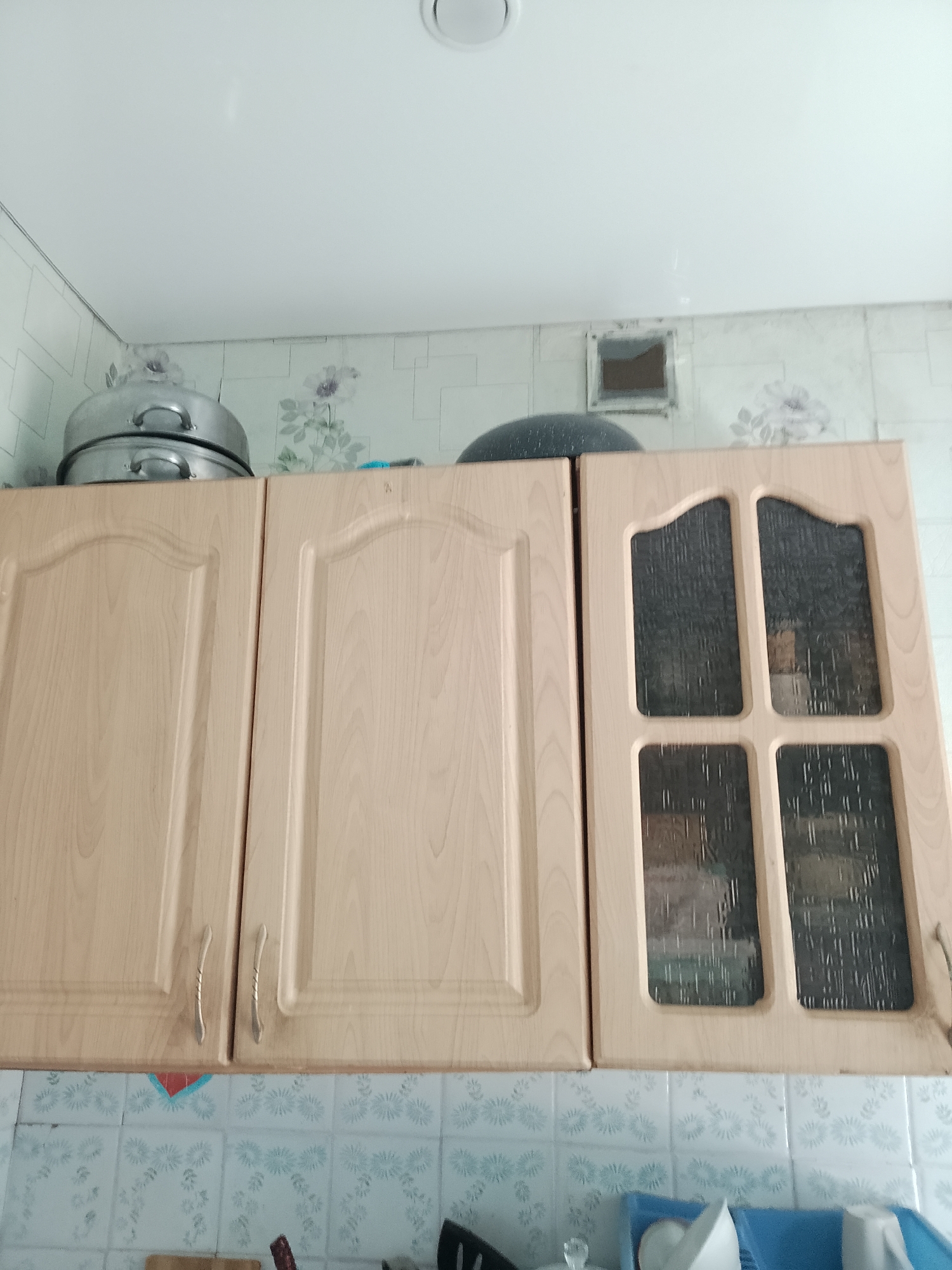 This is a low-angle, eye-level, full shot of kitchen cabinets in the city of Abay, Kazakhstan. The cabinets are made of light-colored wood and have silver handles. The cabinets on the left are solid, while the cabinet on the right has glass doors with a textured pattern. On top of the cabinets are stacked metal pots and pans, and a dark, round object. The wall behind the cabinets is covered in patterned wallpaper with a floral design. Below the cabinets are tiled surfaces. The lighting in the image appears to be artificial, casting a soft glow. The overall scene depicts a functional kitchen environment.