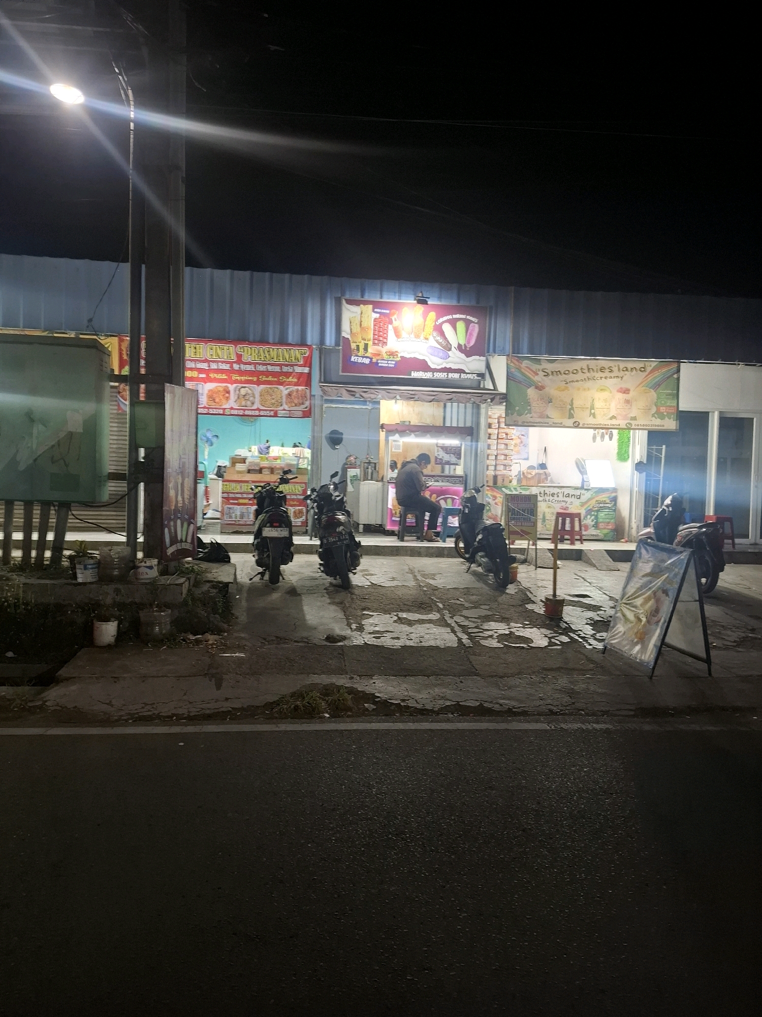 The image depicts a nighttime scene outside a row of small shops in the city of Karangtengah, Indonesia. In the foreground, a paved road is visible, with a sidewalk adjacent to it. Several motorcycles are parked on the sidewalk.
The shops appear to be food vendors. One shop has a banner that reads "Smoothies land" and displays images of smoothies. Another shop has a banner with text that includes "TEH CINTA 'PRASMANAN'" and images of food items. A third shop has a banner that says "WARUNG SOSIS BOH KUNIS".
A person is seated on a stool inside one of the shops, appearing to be working. There are various items displayed for sale, including what appear to be food items and drinks. The shops are illuminated by artificial lighting, and a streetlamp is visible in the upper left corner of the image. The overall environment suggests a bustling commercial area operating into the night.