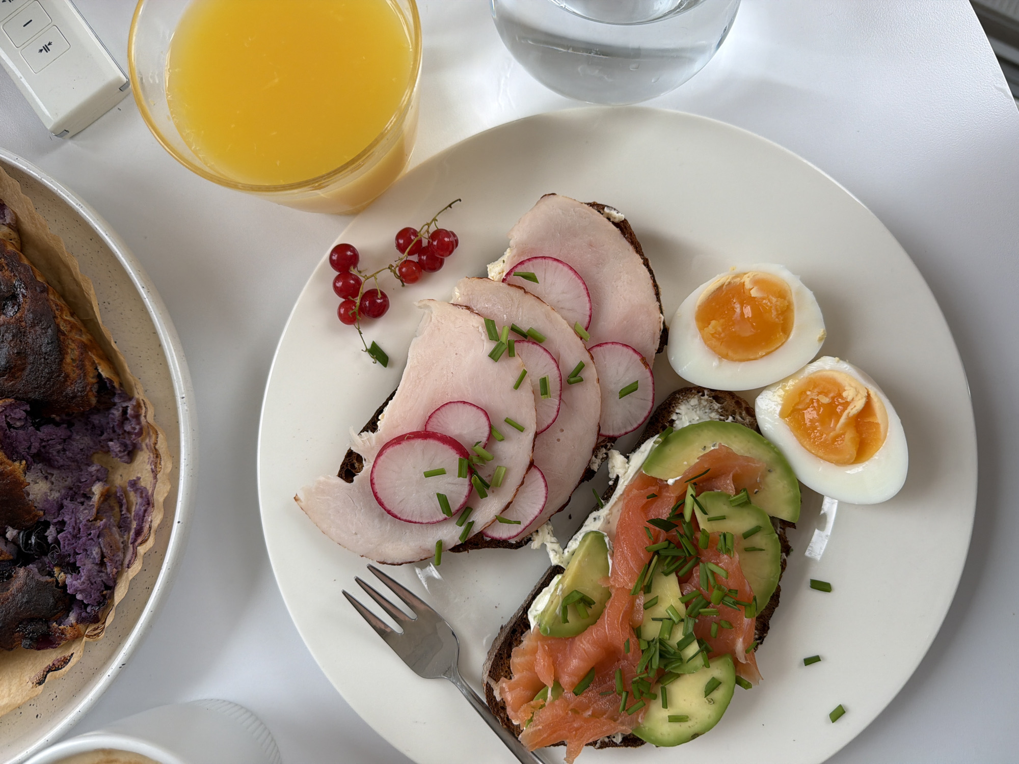 The image depicts a breakfast spread on a white plate, set against a white surface. The plate holds two open-faced sandwiches, one topped with smoked salmon, avocado, cream cheese, and chives, and the other with thinly sliced ham and radishes, also garnished with chives. Two halves of a soft-boiled egg with bright orange yolks are positioned below the sandwiches. To the right of the plate, a glass of orange juice is visible, along with a small cluster of red currants. In the background, partially obscured, is a bowl containing a dark, rustic pastry with visible purple filling and a white cup. A metallic fork rests on the left side of the plate, and a remote control is situated in the upper right corner. The scene appears to be an indoor setting, likely a kitchen or dining area, with no indicators of the specific location within Gangloff, Germany.