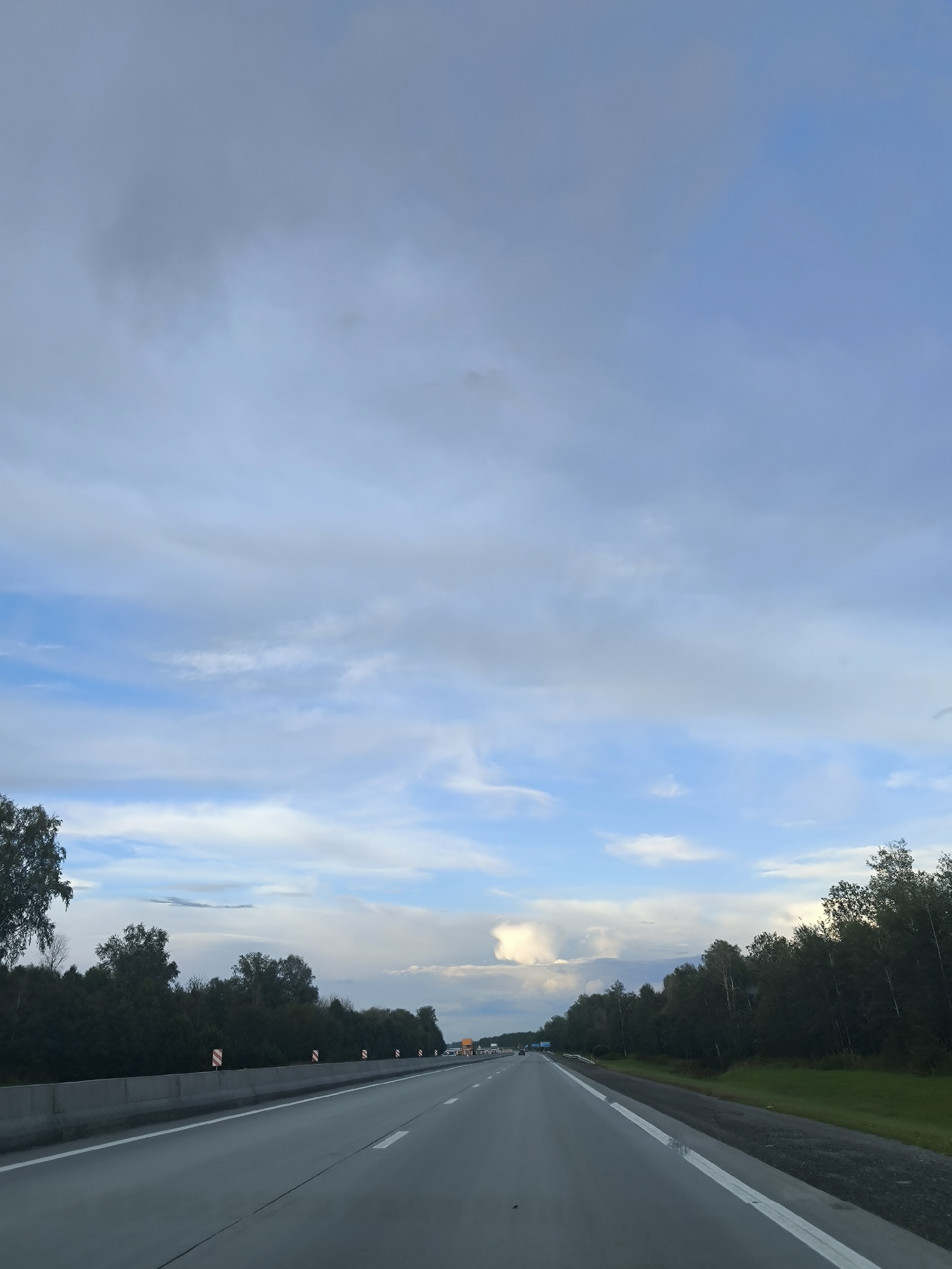 The image shows a view from a vehicle traveling on a highway, likely in or near Sokur, Russia. The highway is paved and has white lane markings. On the left side of the highway, there is a concrete barrier with some warning signs. Beyond the barrier, there is a dense line of dark green trees. On the right side of the highway, there is also a line of trees and a grassy area. The sky is partly cloudy, with a mix of blue sky and grey clouds. There are a few vehicles visible on the highway in the distance.
