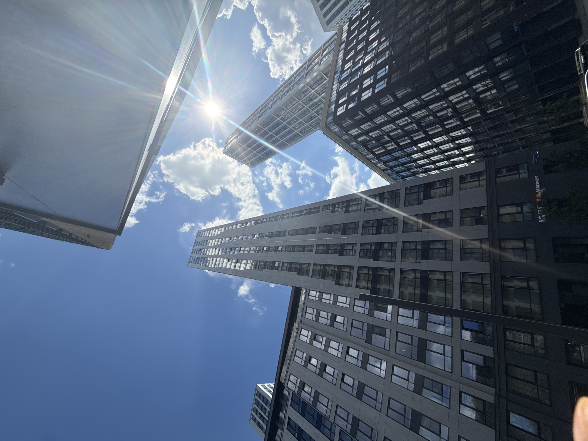 This photograph captures a dramatic worm's-eye view of modern skyscrapers against a bright blue sky with scattered white clouds. The sun is prominently featured in the upper left quadrant, casting a bright flare and lens refraction across the image, indicating a sunny day, likely midday or early afternoon.
The scene is dominated by several multi-story buildings with contemporary architectural designs. A distinctive building in the center features a prominent cantilevered or bridge-like section, connecting different parts of the structure or extending outwards. To the right, another building is darker in color, with numerous reflective windows. A tall, slender building is visible towards the left.
No specific human activity is depicted; the focus is on the urban landscape and architectural forms. The upward perspective emphasizes the impressive height and intricate details of the buildings.
Notable visible text includes a sign on the dark building on the right side of the image. The Cyrillic script clearly reads "Я.Маркет" (Ya.Market), identifying it as Yandex.Market. Below this, a street sign is visible, stating "Пресненская наб. 11" (Presnenskaya nab. 11), which translates to Presnenskaya Embankment 11. These details place the location in Moscow, Russia.