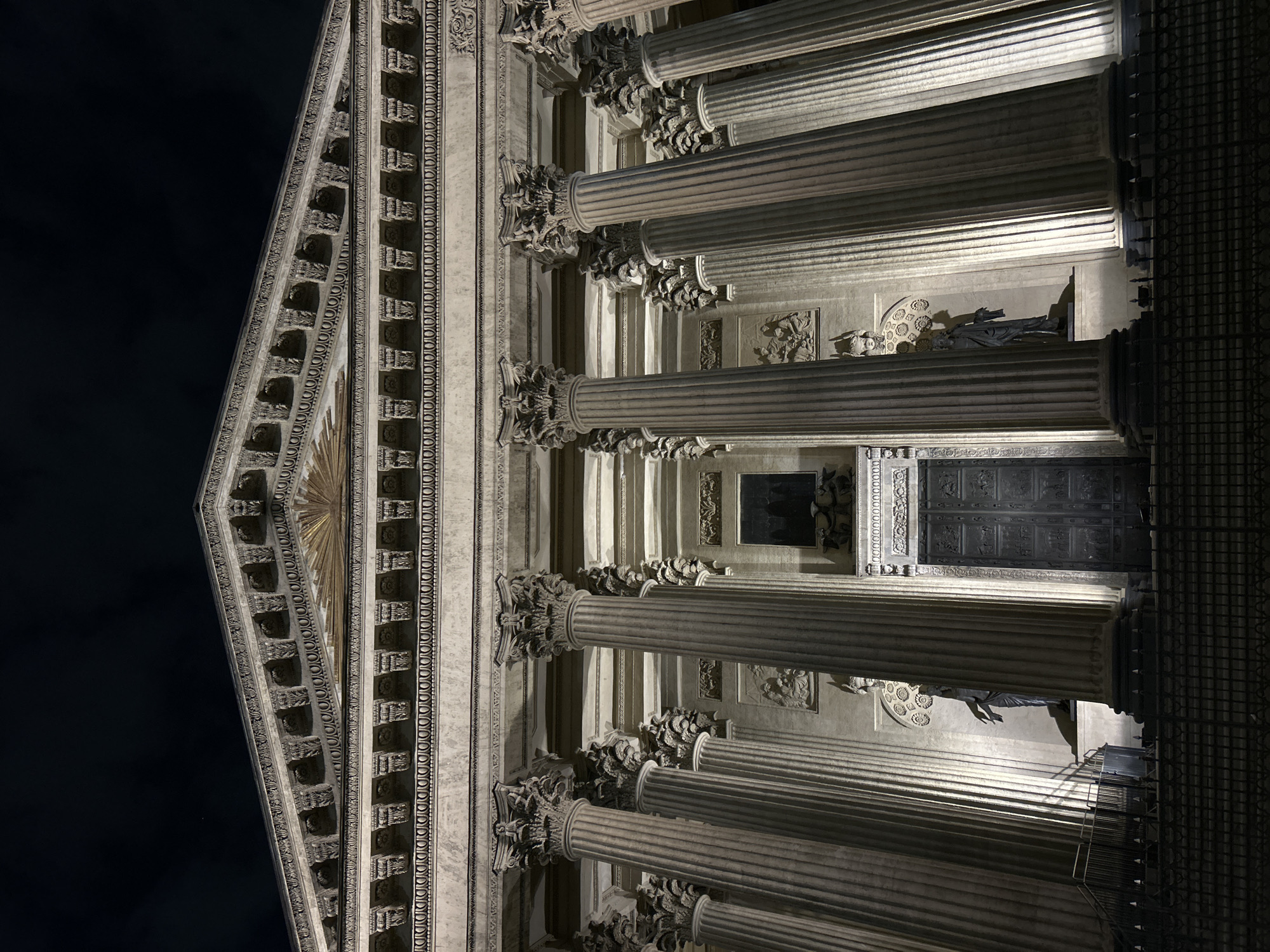 The image is a nighttime, low-angle photograph of the facade of Saint Isaac's Cathedral in Saint Petersburg, Russia. The building's architectural features are prominently displayed, including rows of fluted columns with Corinthian capitals. The triangular pediment at the top of the facade is adorned with sculptural details and a central radiating sunburst design. A large, ornate door is visible between the columns, with relief sculptures on the wall above it. The lighting emphasizes the texture and three-dimensionality of the architectural elements. The sky is a dark, unilluminated background. There are some sculptural figures on a ledge between the columns to the left of the door. A dark railing can be seen towards the right edge of the image.