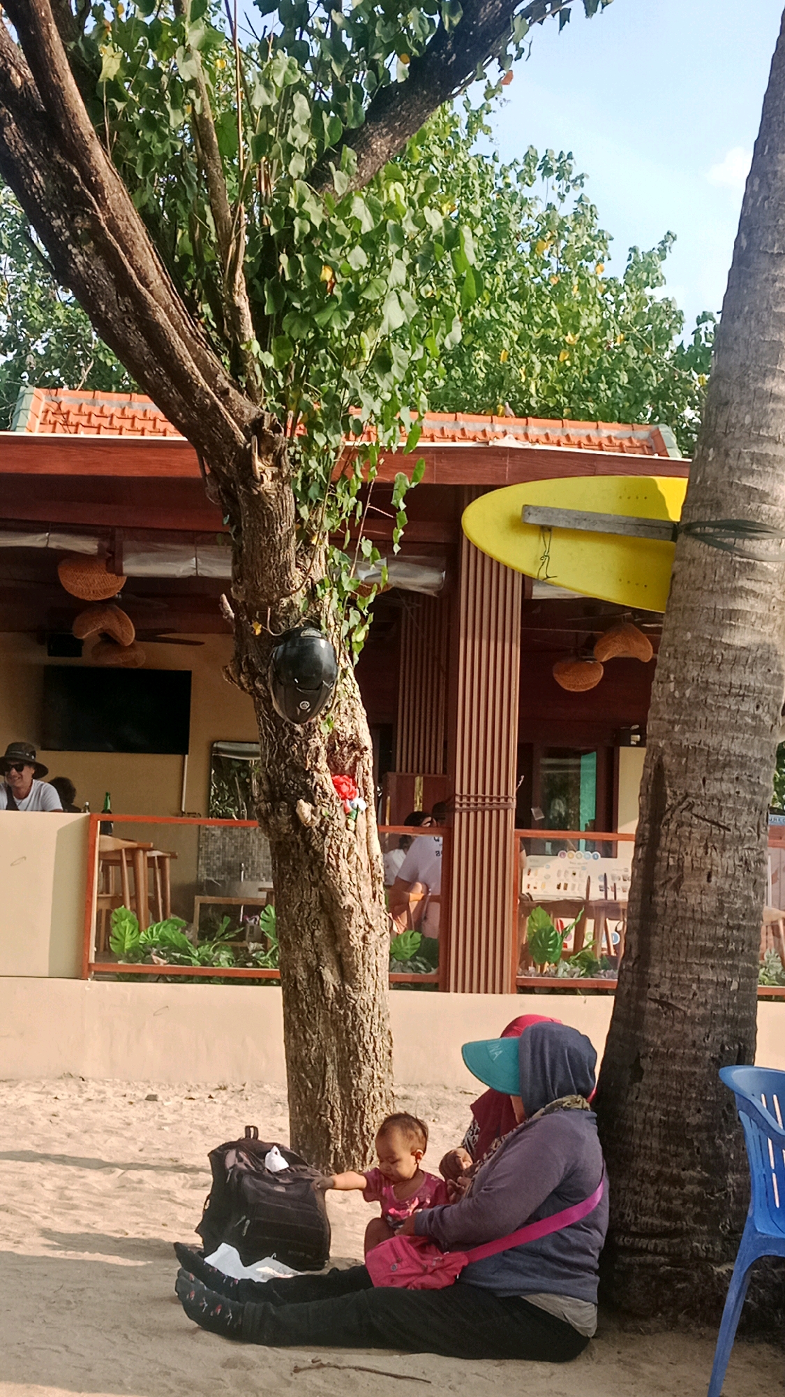 In Kuta, Indonesia, a woman and a baby sit on a sandy beach near a tree and palm trees. The woman, wearing a headscarf and a blue visor, is holding the baby, who is reaching towards a backpack. Behind them, a building with a brown roof and vertical wooden siding is visible, with a yellow surfboard mounted on the exterior. Other people and chairs are also present in the background, suggesting a public area or establishment. The scene is bathed in daylight.