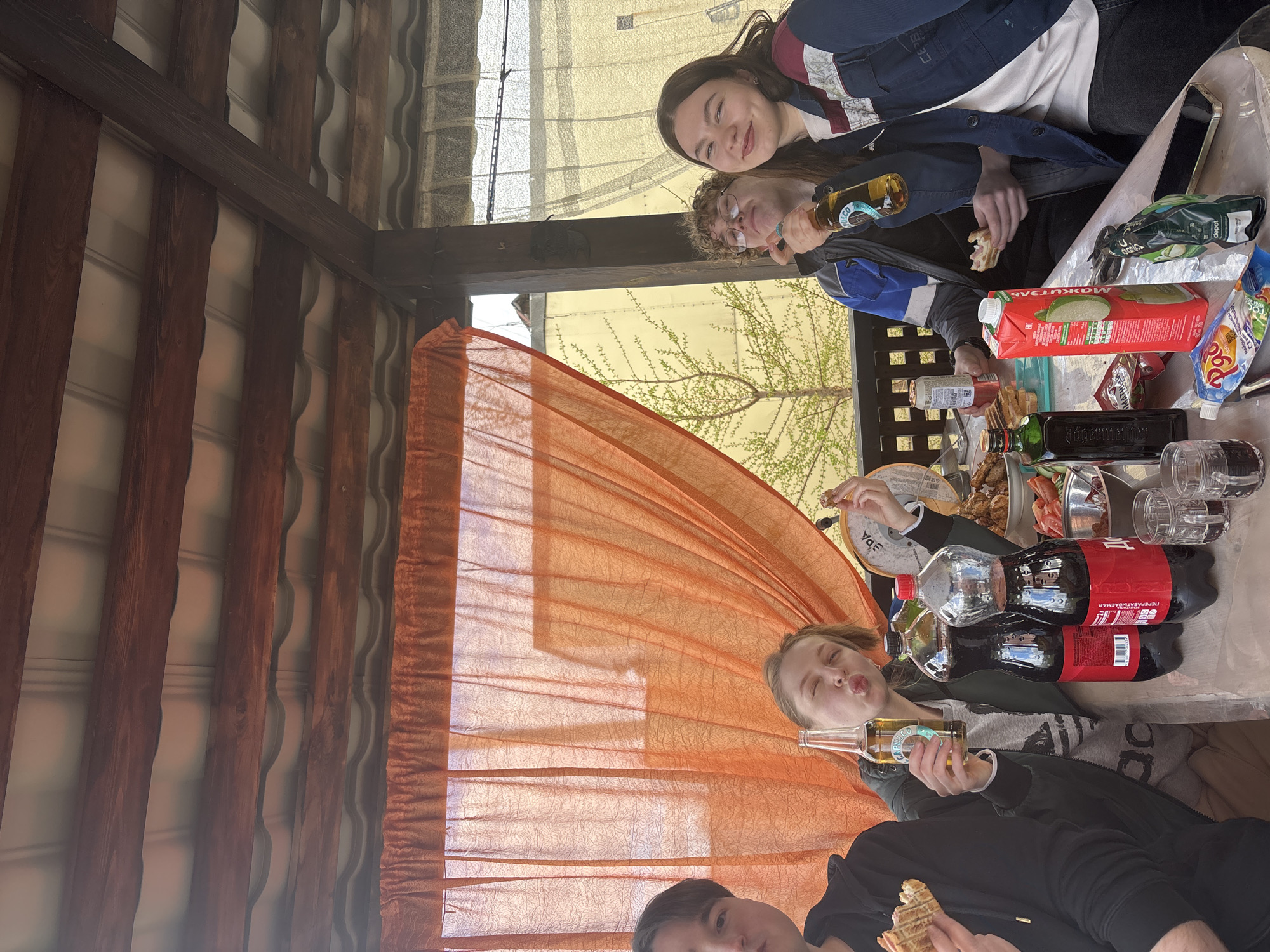 People are gathered at a table under a wooden structure, possibly a gazebo or covered patio. An orange translucent fabric hangs to the side. On the table are food items such as sandwiches, snacks, and drinks, including apple juice, a bottle of Jägermeister, and cola bottles. Several individuals are visible: One person is holding a bottle of beer and making a pouting face, while another person holds a sandwich. A second pair is seen also holding beer and enjoying the meal. One person, a girl, is leaning on the second person.
The setting is in Chaikovsky, Russia. Visible outside the structure are trees and a light-colored building.