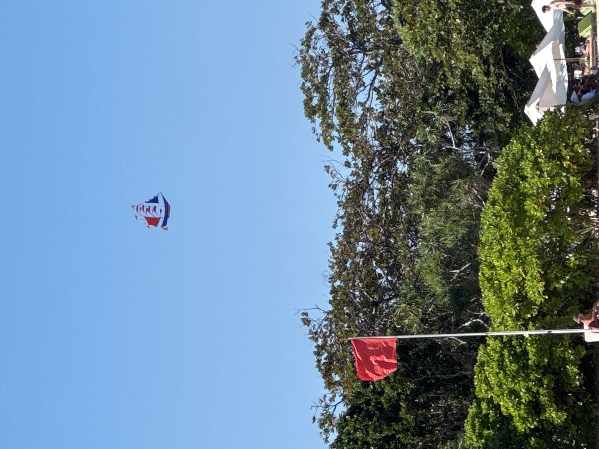 This image depicts a clear blue sky above a lush green treeline. A sailboat-shaped kite, predominantly red and blue with white accents, is flying high in the sky. In the foreground, a red flag waves from a flagpole against a backdrop of dense foliage. Further down, several white beach umbrellas are set up, with a few people relaxing on lounge chairs and walking on the beach. This scene is characteristic of Kuta Selatan, Indonesia, known for its popular beaches and recreational activities.