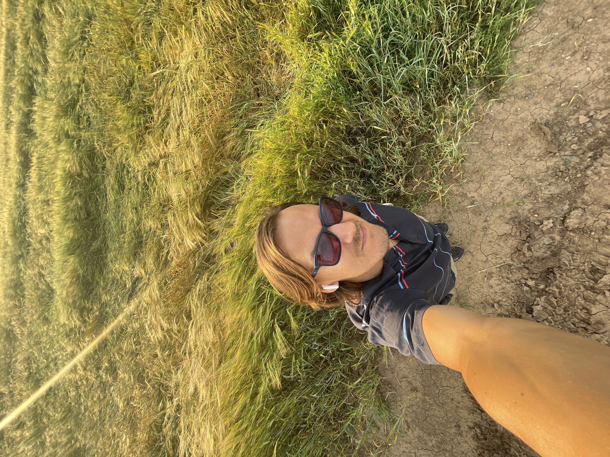 An adult male, with medium-length brown hair, is taking a selfie in a field of tall grass. He is wearing sunglasses and earbuds. The sun is low in the sky, casting a strong glare across the field. The ground is dry and cracked, suggesting arid conditions. The location is the village of Ol'khovo, Russia.