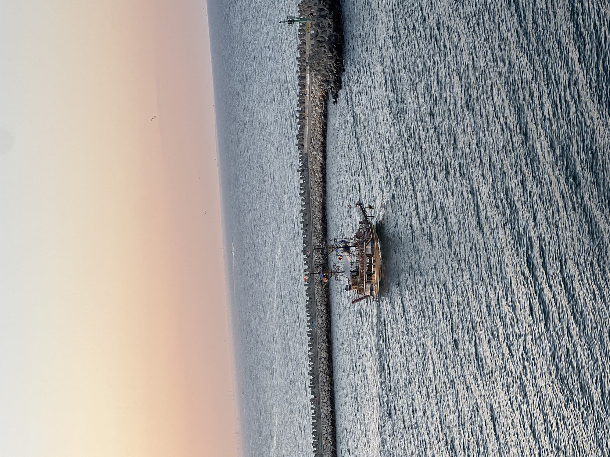 The image captures a pirate-themed boat sailing on the Black Sea near the Constanța Harbor, Romania. The scene is set during sunset, with a pastel-colored sky transitioning from pale yellow to soft pink. A breakwater, constructed of concrete tetrapods, stretches across the water, guiding ships into or out of the harbor. A green lighthouse stands at the end of the breakwater, signaling the entrance. The pirate boat, adorned with flags and a wooden exterior, carries passengers who appear to be enjoying the twilight cruise. The water exhibits gentle ripples, reflecting the serene ambiance of the evening. The distant shoreline suggests an urban or industrial landscape characteristic of a port city.