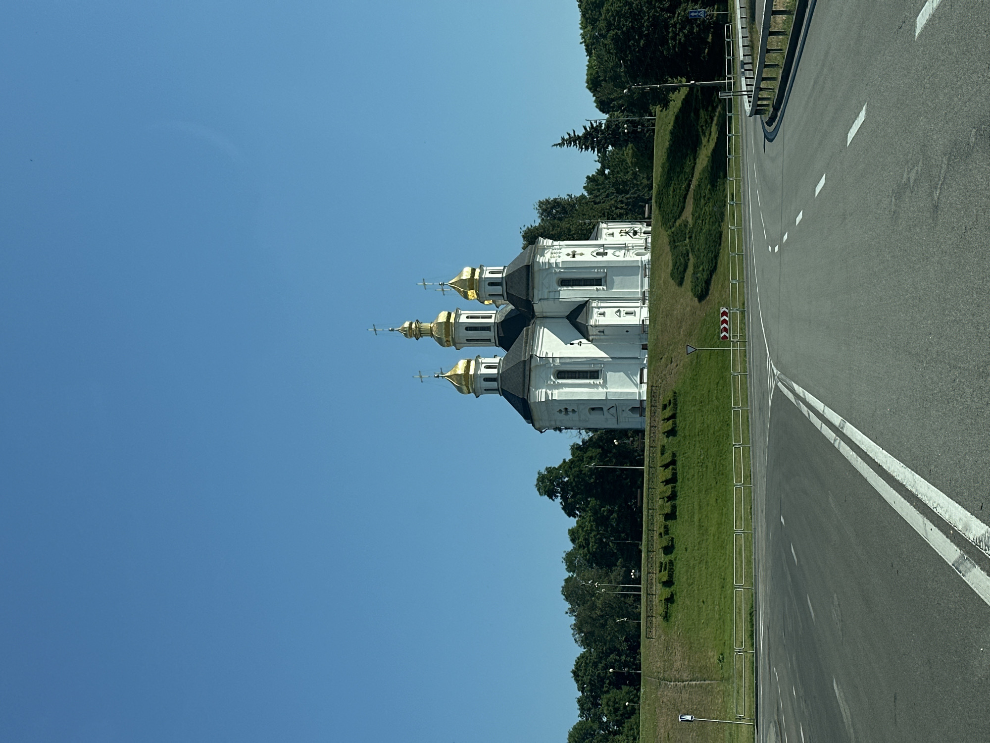 This is a photograph taken from a street level perspective, looking towards a prominent religious building. The building is a white church with multiple golden domes, each topped with a cross. The church is situated on a green, grassy hill, with a line of trees forming a backdrop behind it. In front of the hill, there is a road intersection with distinct white lane markings. The sky above is a clear, bright blue. The scene suggests a sunny day in an urban or semi-urban environment, likely Chernihiv, Ukraine, given the distinctive architecture. There are no people or vehicles immediately visible in the frame.
