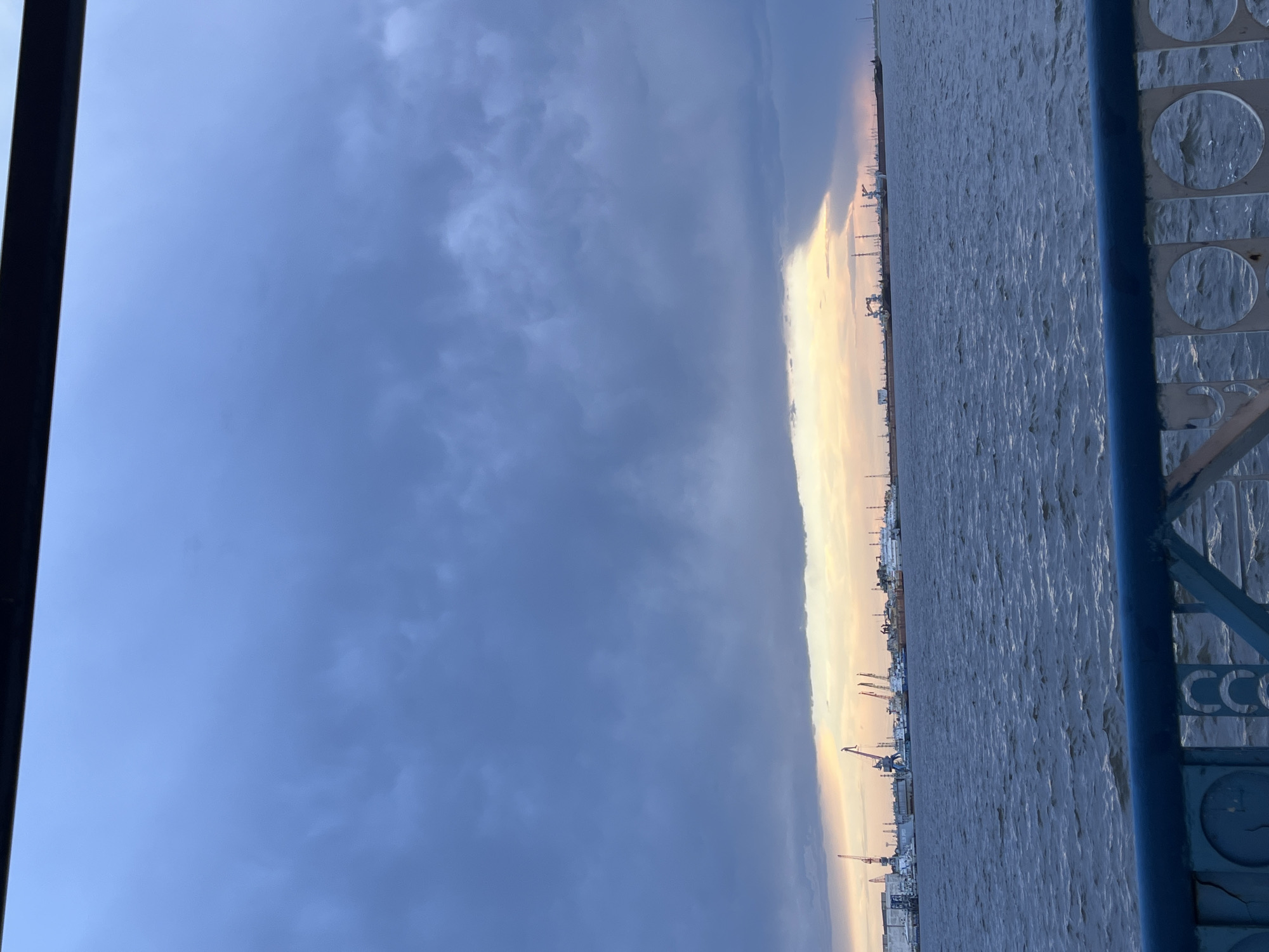 The image shows a view from a high vantage point, possibly a bridge, in Chaikovsky, Russia. The scene is dominated by the Kama River, its surface reflecting the overcast sky. Along the riverbank, there is an industrial area with cranes and various structures. The sky is filled with heavy clouds, partially illuminated by a narrow band of sunlight. On the right side of the image, part of a blue-painted metal railing with decorative elements is visible. The letters "CCC" are visible on it. On the left side of the frame is a black vertical line which may be another structural element of the bridge or viewing point.