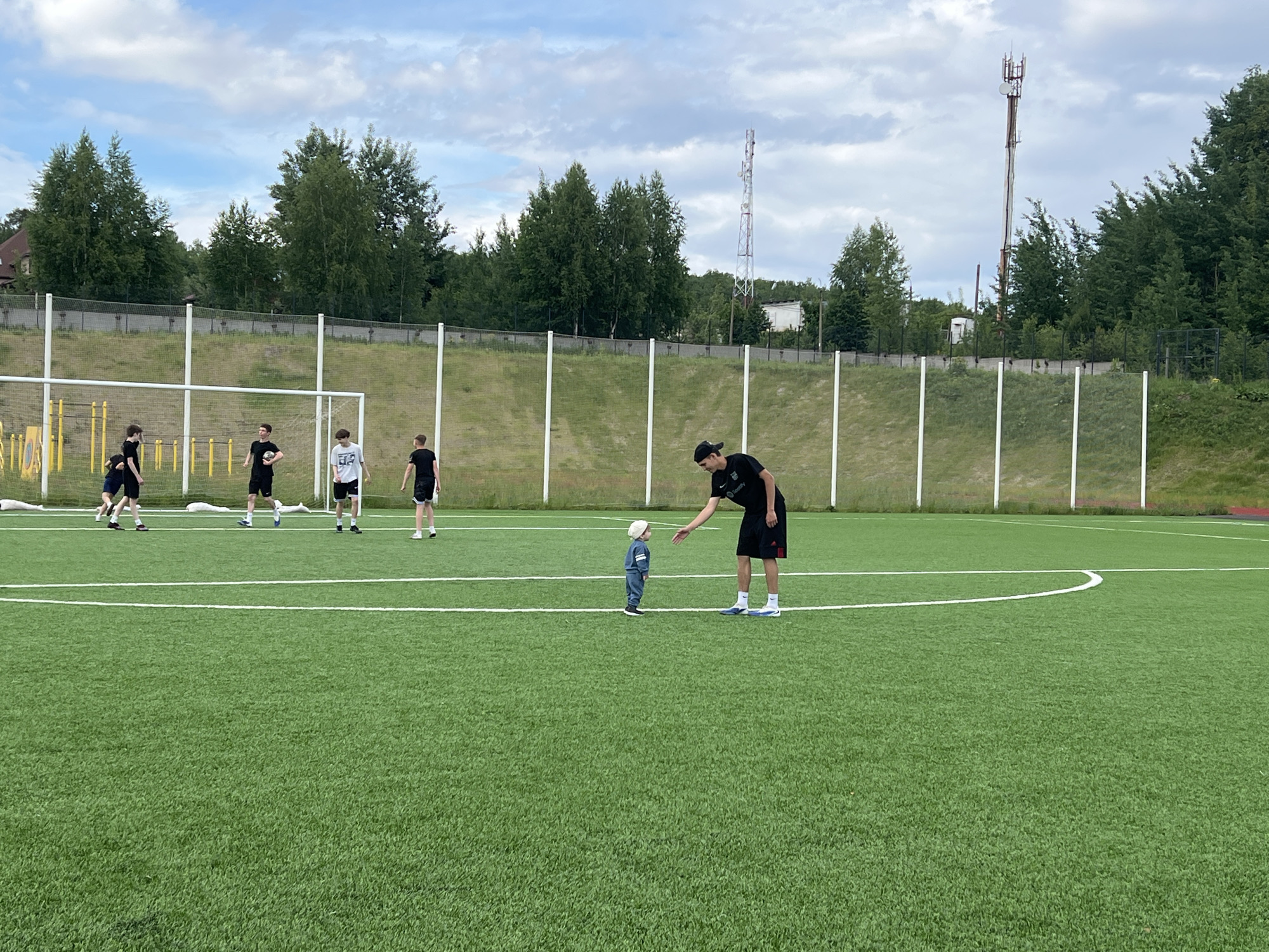 The image shows a group of people on a green soccer field in Noviy, Russia. There are five figures to the left, near a soccer goal, apparently playing or practicing. A man wearing a black shirt and shorts and a black baseball cap is bending slightly, with one arm extended towards a small child standing on the field. The child is wearing a blue outfit and a light-colored hat. The background includes trees, a white fence, and a tower structure. The sky is partially cloudy.