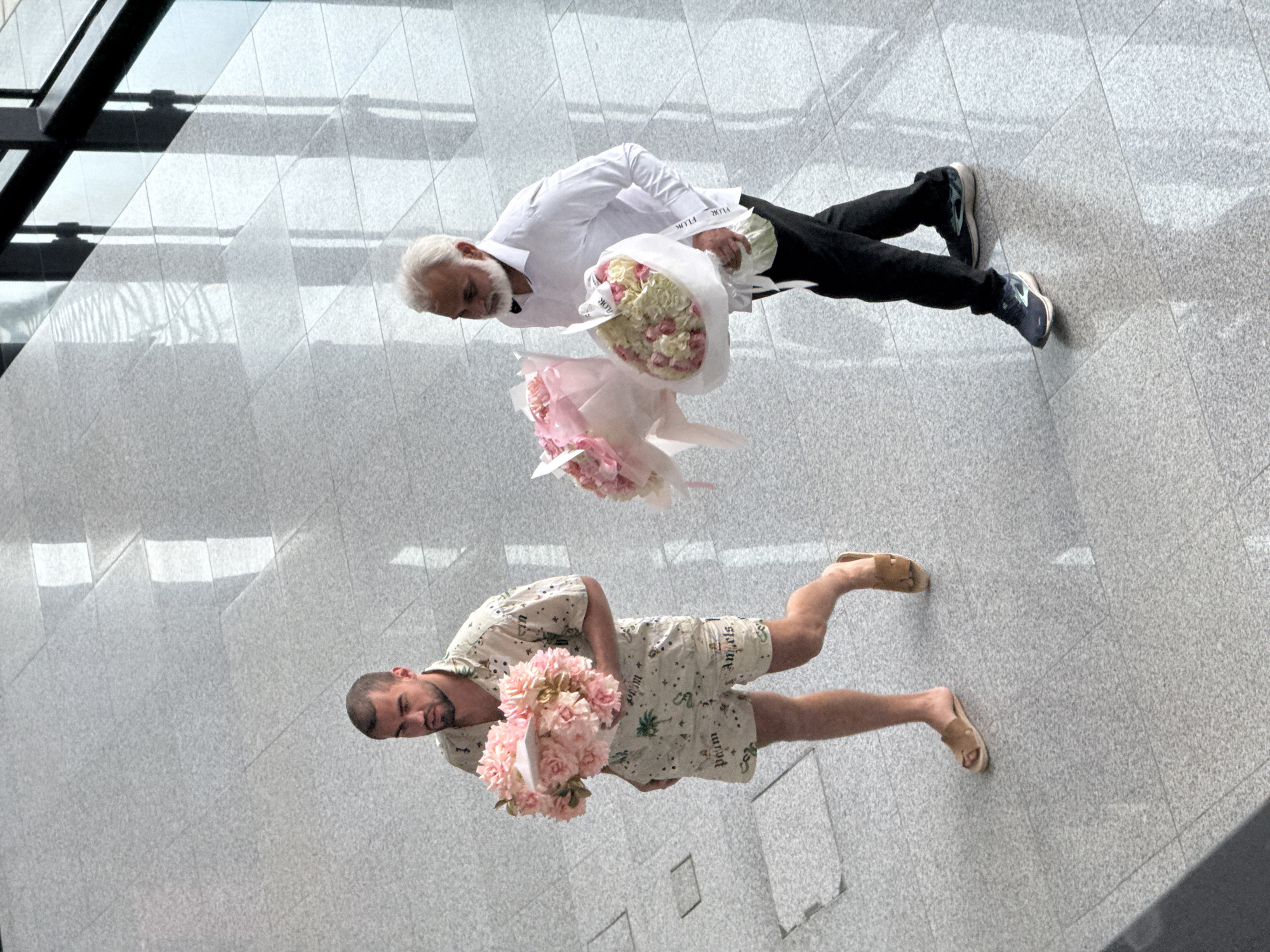 Two men stand on a grey tiled floor in a modern indoor setting, likely a large public space such as a shopping mall or hotel lobby, consistent with a location like Dubai, United Arab Emirates. The bright lighting suggests it is daytime, with reflections from large windows or glass structures visible in the background.
The man on the left, older with white hair and beard, wears a white long-sleeved shirt, black trousers, and sneakers. He holds two bouquets of flowers: one larger bouquet of white and pink roses wrapped in white paper, and a smaller bouquet of light pink flowers wrapped in pink paper, which appears to be suspended as if being passed or caught mid-air. The wrapping of one bouquet clearly shows the word "FLOR".
The man on the right, younger with dark hair and a beard, is dressed casually in a matching patterned short-sleeved shirt and shorts with visible "Palm Angels" and "Angels" branding. He wears light-colored sandals and holds a large bouquet of pink flowers.
Both men are looking down towards the flowers they are holding, suggesting they are either admiring them, posing for a photo, or preparing to present them. The overall scene conveys a calm atmosphere.