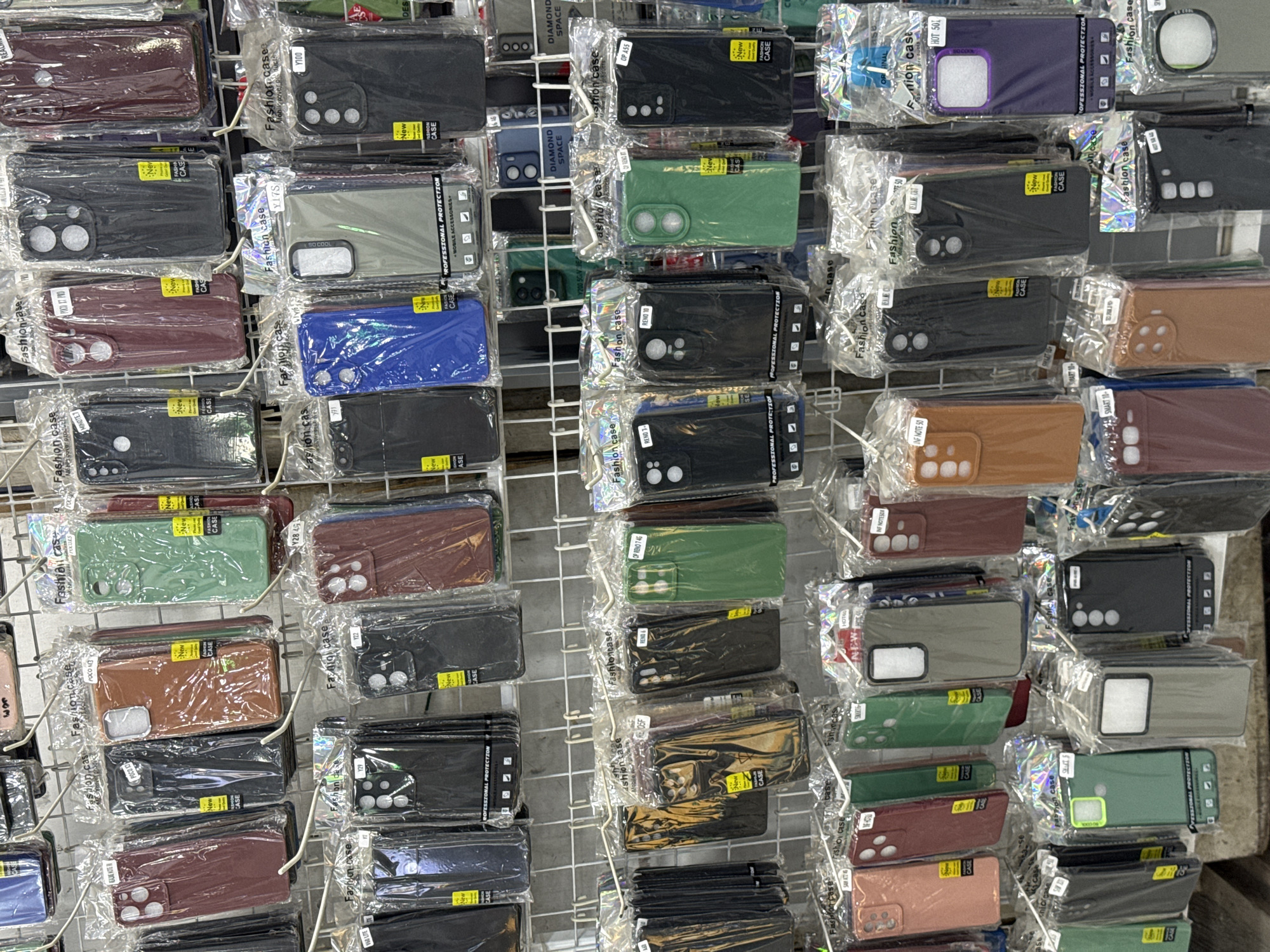 The image displays a retail display of mobile phone cases arranged on wire racks. The cases are packaged in clear plastic and feature various colors and designs, with some indicating specific phone models. The background consists of a plain wall. The image is cropped from a larger scene, and the provided crops offer closer views of different sections of the display. No people are visible in the image. The context suggests a shop or market stall selling phone accessories.