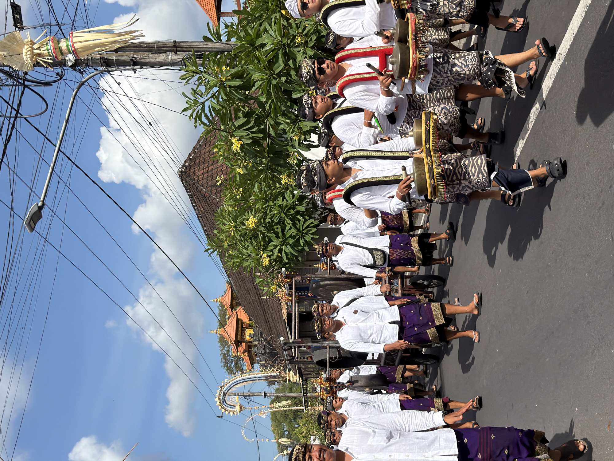 A procession of men in traditional Balinese attire walks down a paved street under a bright, cloudy sky in Mengwi, Indonesia. The men are wearing white long-sleeved shirts, purple sarongs with gold trim, and headbands. Many are playing musical instruments, possibly gamelan, consisting of metal gongs and cymbals arranged on stands. Some men are pushing a cart decorated with intricate carvings and fabric. In the background, traditional Balinese architecture with tiered roofs and ornate gateways can be seen, surrounded by lush green trees. Overhead, a network of power lines and a street lamp with a decorative banner are visible. The overall scene depicts a cultural event or ceremony taking place in a village setting.