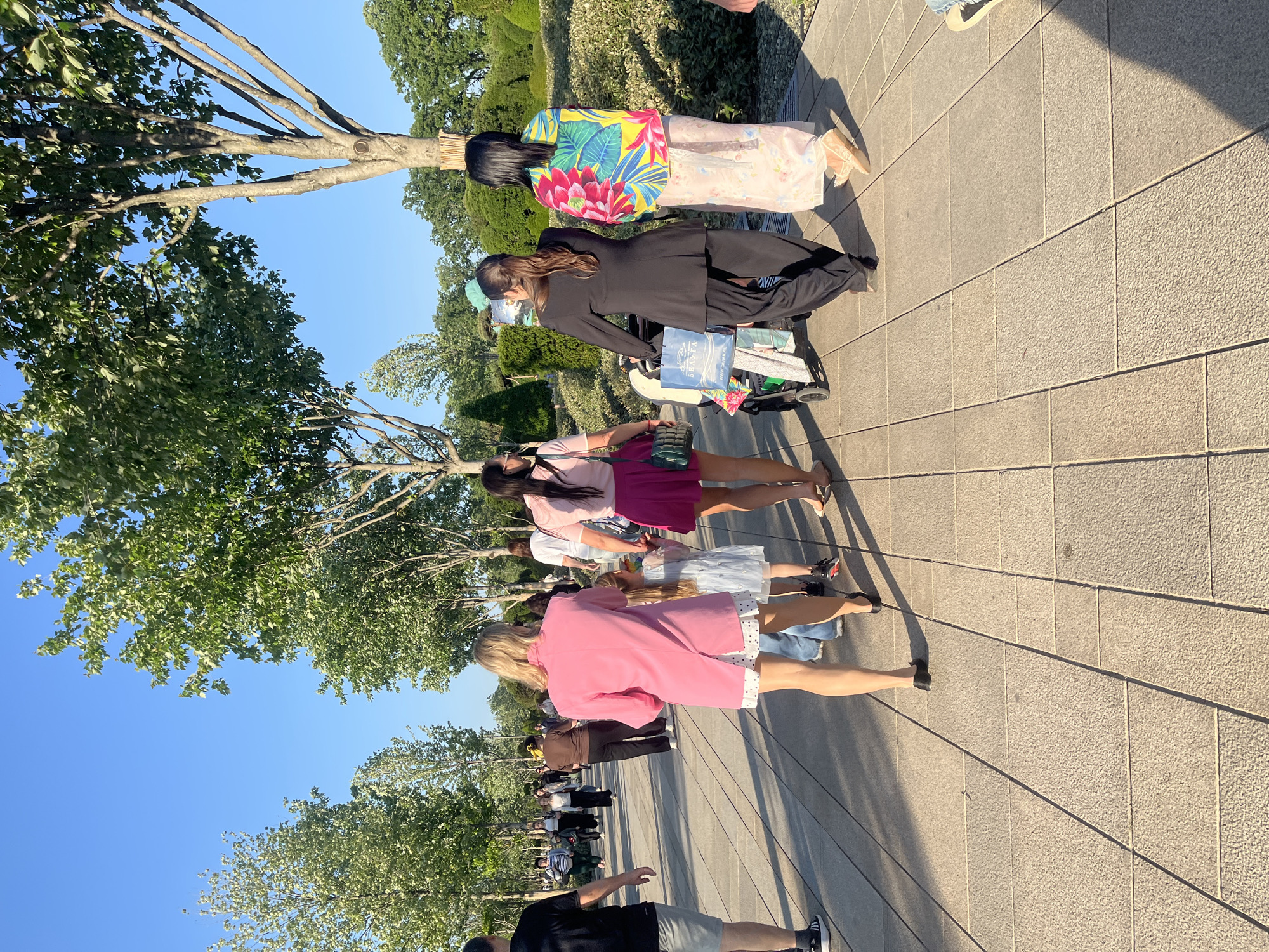 The image shows a group of women walking along a tiled pathway. The women are dressed in various styles of clothing, including casual dresses, a pink blazer, a black suit, and a floral kimono. One woman is pushing a baby stroller. Trees line the walkway. The sky is blue and clear. The location is likely a public park or plaza in Krasnodar, Russia.