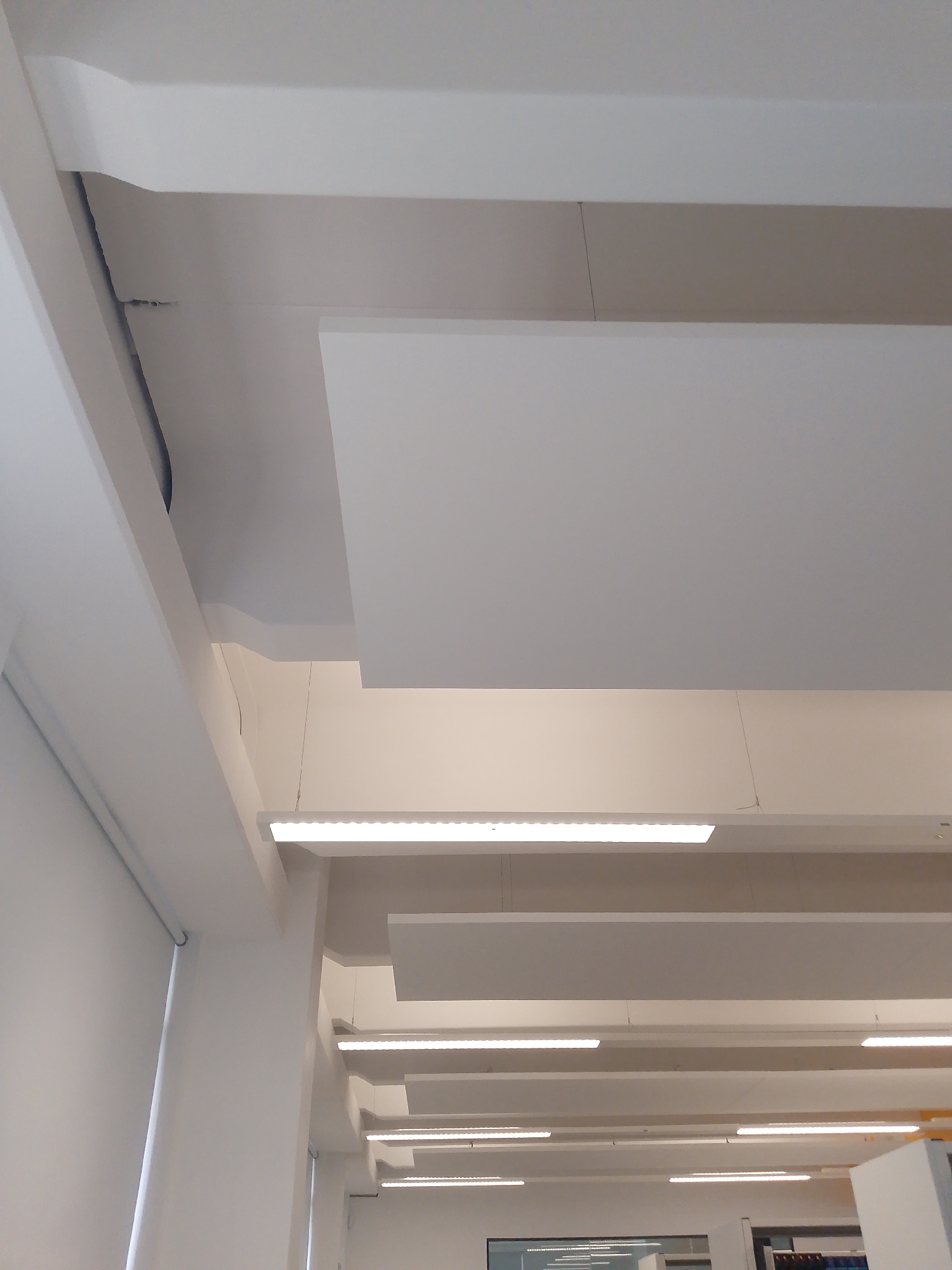 This image offers an upward view of a modern, brightly lit interior space, likely an office, library, or institutional building in Coventry, United Kingdom. The predominant colors are white and various shades of light grey, contributing to a clean and minimalist aesthetic.
The ceiling features a multi-layered design with several prominent white architectural beams running across the frame. Suspended beneath the main ceiling structure are numerous rectangular white acoustic panels, arranged in parallel rows at different depths. These panels are held by thin, almost invisible wires. Interspersed between and below these panels are long, linear LED strip lights, providing bright and even illumination to the area. Some of these lights appear integrated into the ceiling structure, while others are suspended.
To the upper left, a dark pipe or cable runs along a recessed channel in the ceiling, hinting at the building's infrastructure. On the far left, a white wall is partially visible, featuring a neatly rolled-up white window blind. The overall lighting suggests it is daytime, or the space is entirely lit by artificial sources, creating a uniformly bright environment.
No people are visible, and there is no discernible activity taking place, emphasizing the architectural and design elements of the room. The bottom right corner of the last cropped image offers a slight glimpse of furniture or room partitions, confirming it as an indoor, functional space. No readable text is visible anywhere in the images. The design emphasizes functionality, sound dampening, and modern aesthetics.