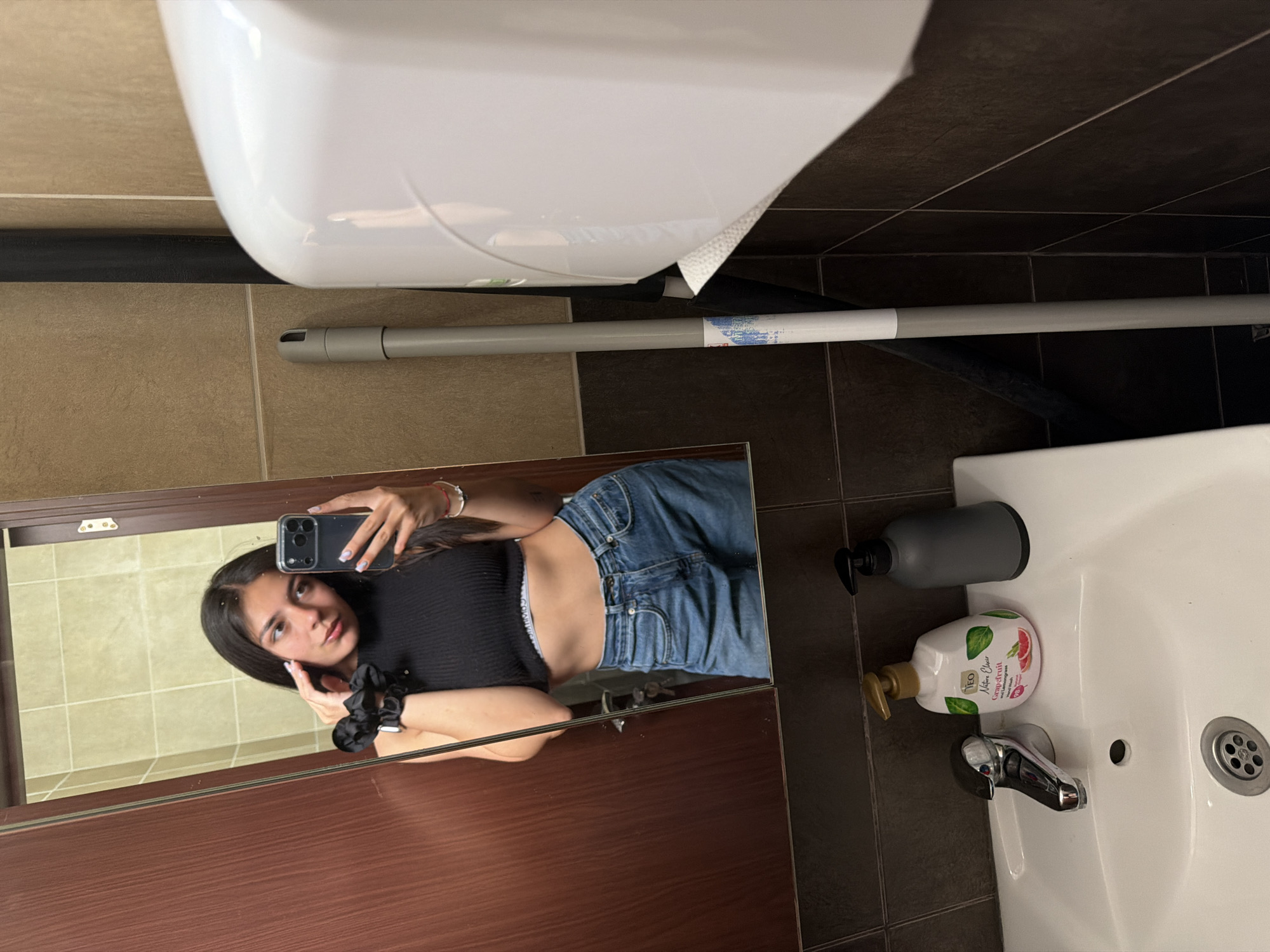 A young woman takes a mirror selfie in what appears to be a bathroom in Gabrovo, Bulgaria. The image is rotated ninety degrees counter-clockwise.
The woman, with long dark hair, is reflected in a large mirror framed by dark wood. She is wearing a black ribbed crop top and blue jeans, with the waistband of "Calvin Klein" underwear visible above her jeans. She holds a smartphone with a black case to capture the image, looking directly at the phone. A black scrunchie is on her left wrist, and her nails appear manicured.
The bathroom features tiled walls, with lighter beige tiles reflected behind the woman and darker brown tiles around the sink and hand dryer area. A white ceramic sink with a chrome faucet is visible to the right. On the sink's edge or counter, there are two soap dispensers: one dark grey and one white. The white dispenser has text that reads "NEO Nature Clear Grapefruit Hand Wash". Above the sink area, a white automatic hand dryer is mounted on the wall. A grey mop handle with a blue and white label (text unreadable) is resting against the dark tiled wall, and a black toilet brush holder is partially visible below it. The lighting suggests an indoor setting, likely during the day or evening with artificial light.