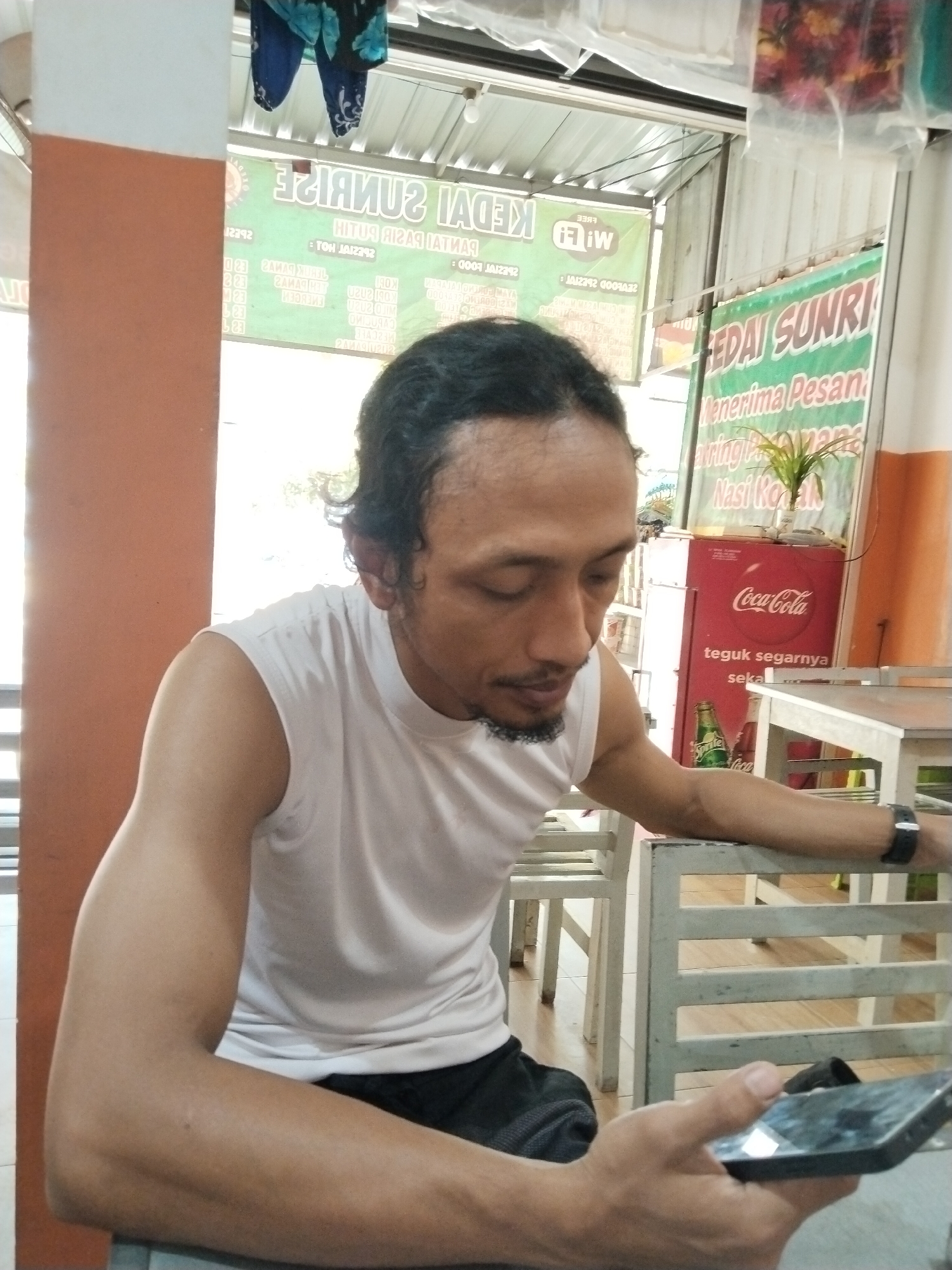 A medium shot captures a man with dark, curly hair and a goatee sitting at a white, slatted table, looking down at a black smartphone he holds in his right hand. He wears a white sleeveless t-shirt and black shorts. His left arm rests on the back of a chair. Behind him, several signs are visible: one with the text "KEDAI SUNRI" in green and white, and another with a Coca-Cola logo on a red refrigerator. A potted plant sits on the refrigerator. The environment appears to be outdoors, possibly at a roadside stall or restaurant, with a corrugated metal roof overhead and an orange pillar on the left. The lighting suggests it is daytime. The scene takes place in Prigi, Indonesia.