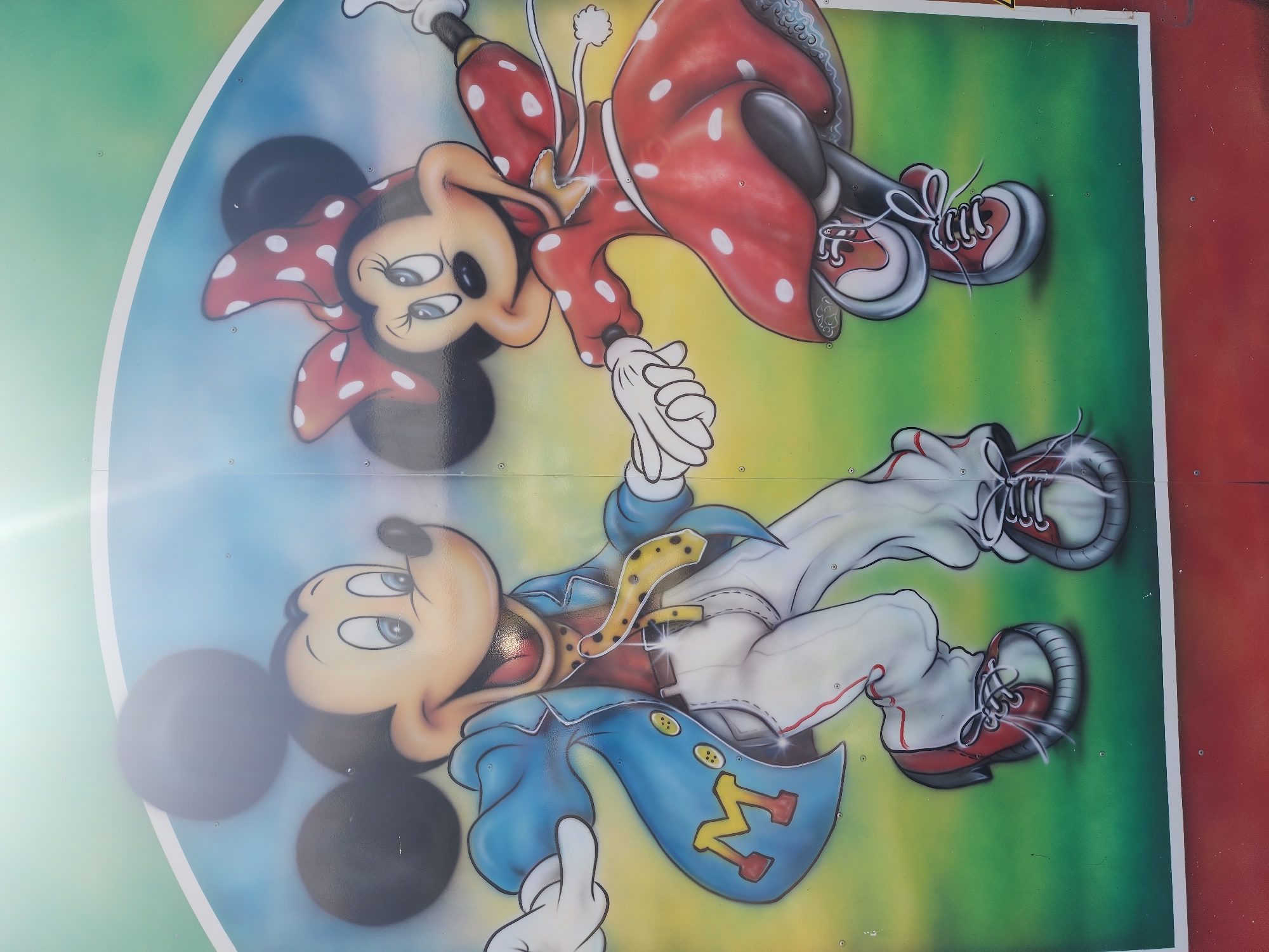 A painted image of Mickey and Minnie Mouse is located in Kazan, Russia. Mickey Mouse is dressed in a blue coat with an "M" on the lapel and white pants. Minnie Mouse is dressed in a red polka dot dress and red shoes. Both characters are wearing sneakers and holding hands. They are painted on a surface that is multicolored and has a white border. The image is outdoor and shows signs of wear.