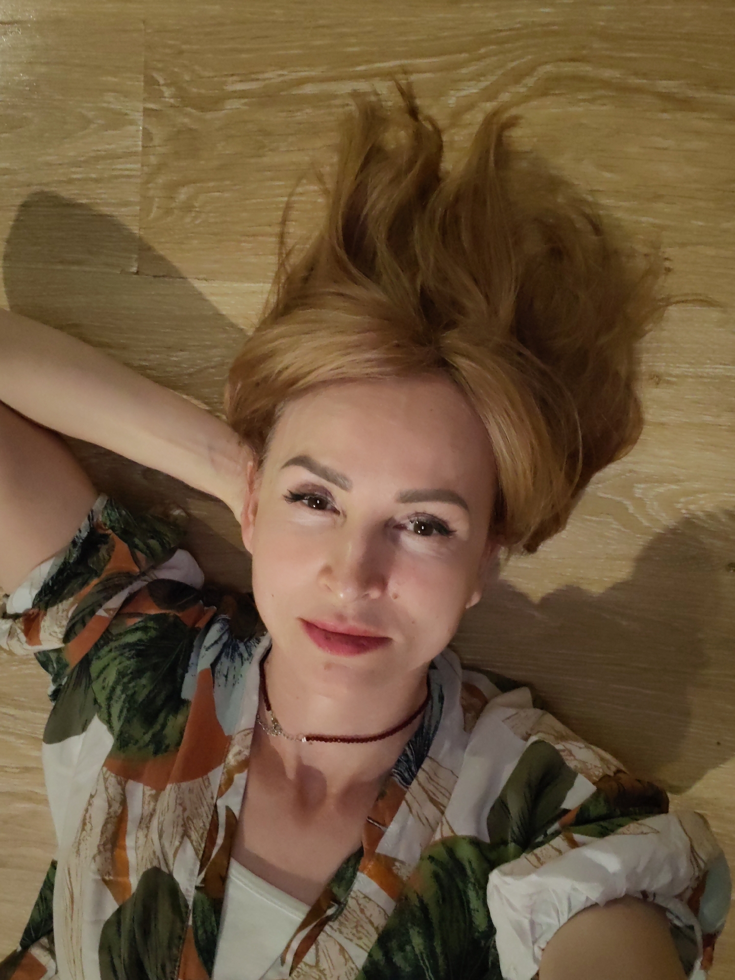 A fair-skinned woman lies on her back on a wood-patterned floor. She is wearing a patterned blouse with white, green, and brown leaf-like designs. A thin, red choker necklace adorns her neck. Her blonde hair is spread out behind her head, and her left arm is bent behind her head, supporting it. Her eyes are open, and she is looking at the camera with a slight smile. A shadow is visible on the floor near her hair. The location is Chaikovsky, Russia.
