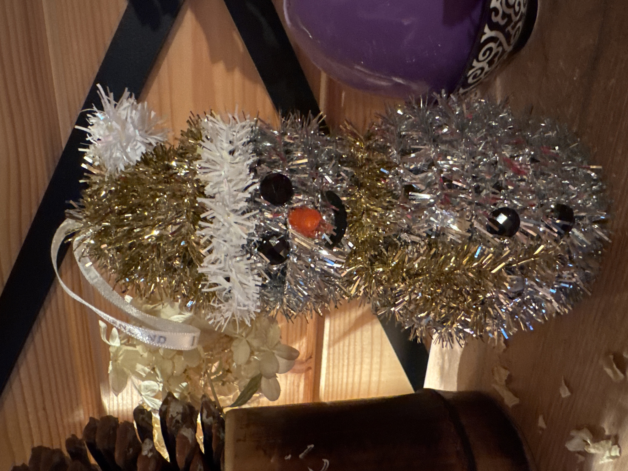 The image shows a close-up of a tinsel snowman ornament. The snowman is decorated with gold and silver tinsel, and has black bead eyes, an orange carrot nose, and a black mouth. It is wearing a white tinsel hat with a white pom-pom. A white ribbon is attached to the hat, presumably for hanging. The snowman is sitting on a wooden surface, with a pine cone and dried flowers to its left, and a purple spherical object with an ornate pattern on its right. The background is a wooden wall. The image does not provide any information about the city of Zelenaya Roshcha, Russia.