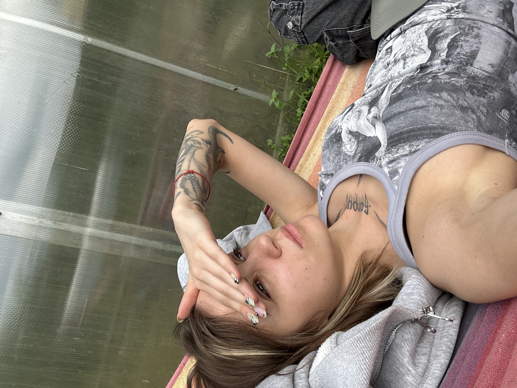 A young person with blonde-streaked brown hair lies in a hammock in what appears to be a greenhouse. The person is wearing a gray tank top with a black and white print, dark jeans, and a gray zip-up hoodie. Their right arm is extended, hand resting on their forehead, as if shielding their eyes from the sun. The person has tattoos on their right arm and neck, and their fingernails are painted with a colorful, abstract design. The background consists of the translucent, corrugated walls of a greenhouse, with glimpses of green foliage and a cloudy sky visible through the panels. The hammock is made of striped fabric in shades of pink, yellow, and red. The overall impression is of a relaxed, outdoor scene, possibly in a rural or semi-rural setting, potentially within the grounds of a dacha or private residence. The location within the city of Козино, Russia, is not explicitly evident from the visual cues in the image.