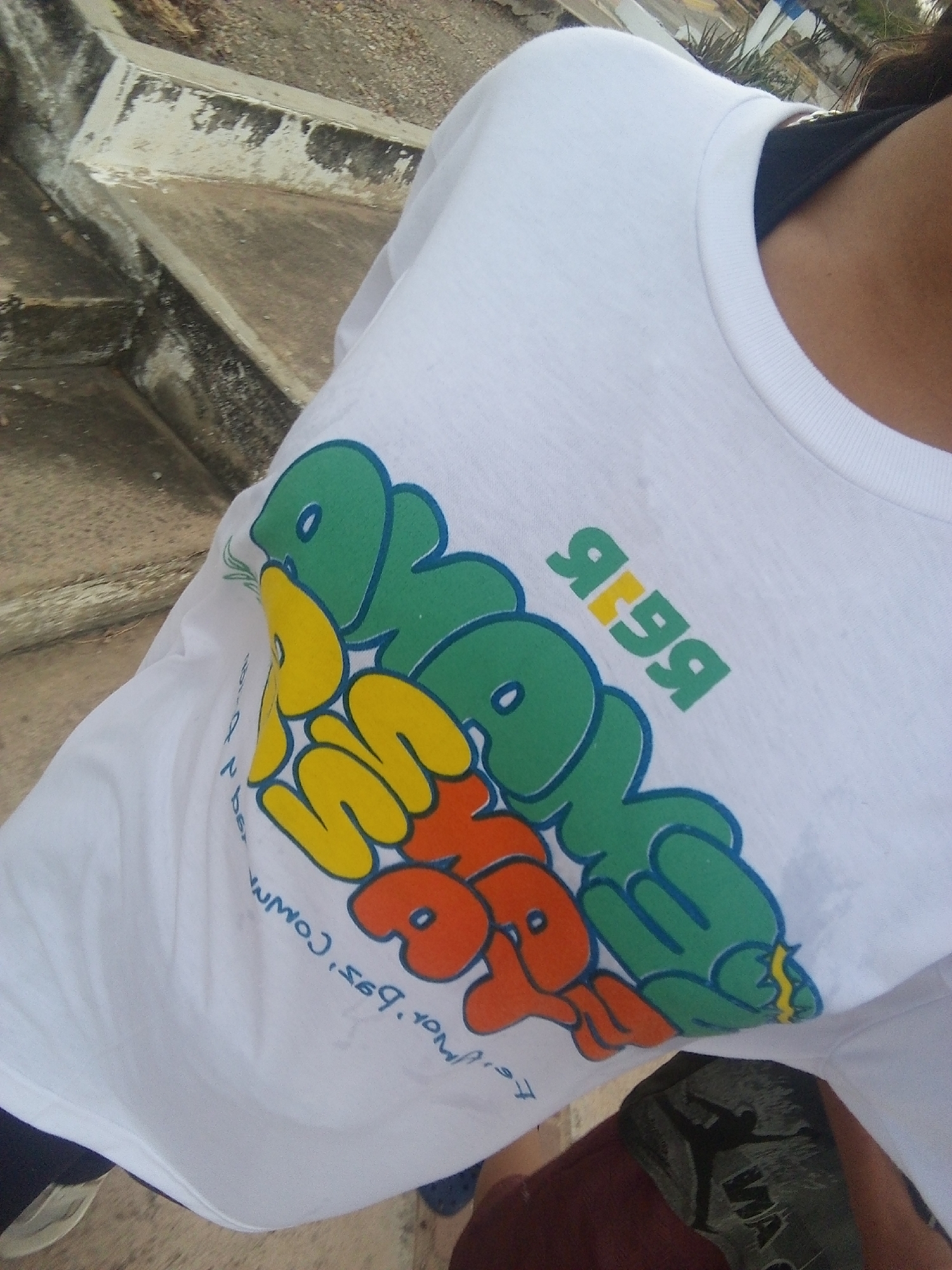 A person wearing a white T-shirt with a colorful graphic is standing on or near concrete steps, likely outdoors. The T-shirt's design features lettering in green, yellow, and orange. The background shows a mix of concrete structures and glimpses of vegetation, suggesting a location in a developed, possibly urban, setting. The details of Juan Griego, Venezuela are not discernible from the image.